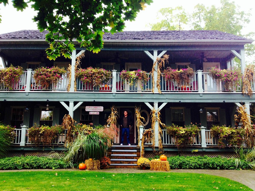 A charming two‑storey inn in Niagara‑on‑the‑Lake with flower boxes adorning the upper balcony, a covered porch beneath, and lush greenery surrounding the building under a clear blue sky