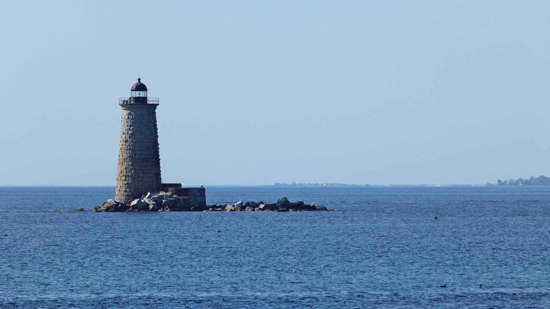 A stone lighthouse, Whaleback Light, stands on a rocky outcrop in the middle of a calm, blue body of water under a clear sky.