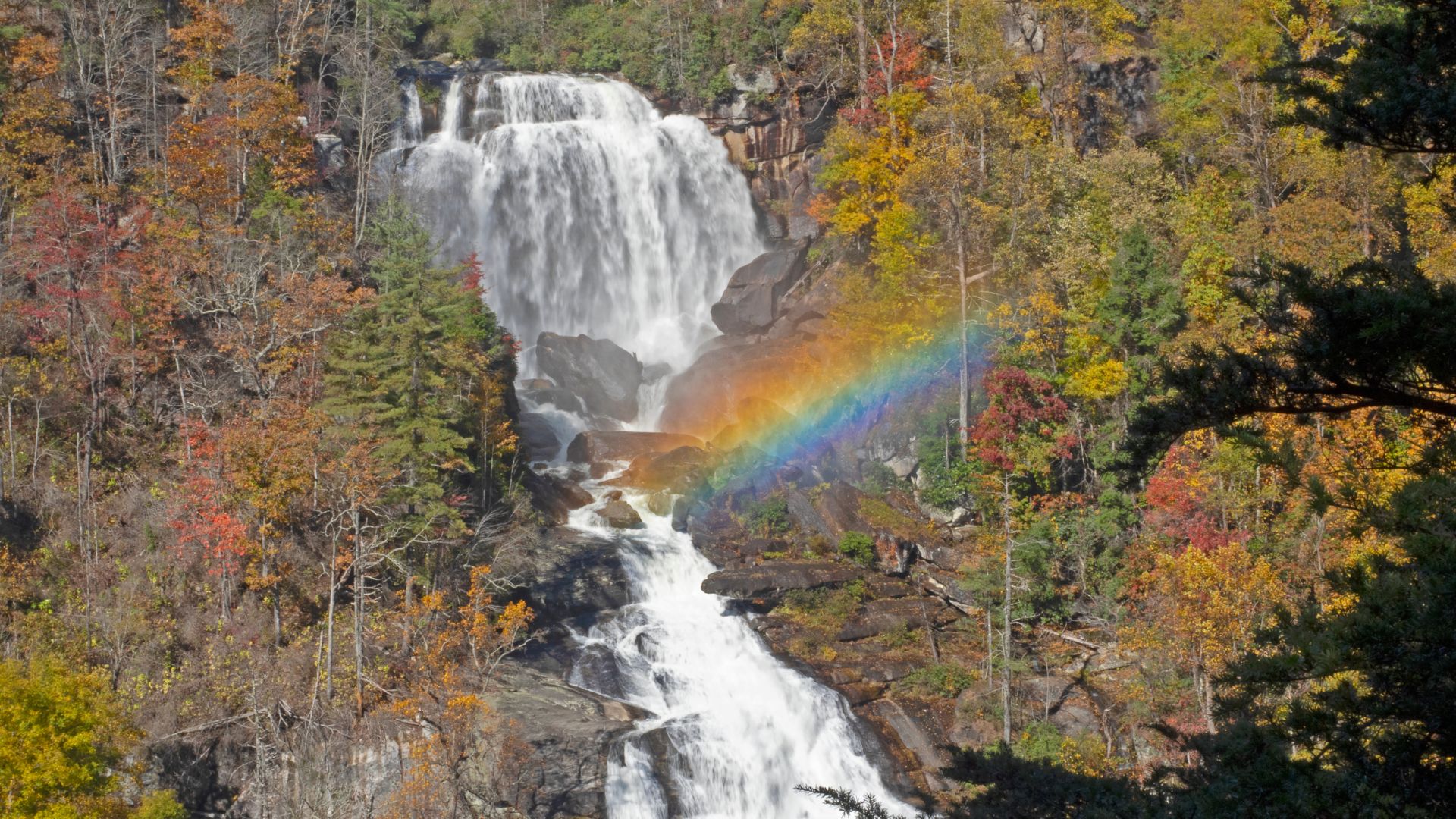 A majestic waterfall cascades over multiple rock tiers amidst vibrant autumn foliage, with a bright rainbow arcing across the mist at the base of the upper falls.