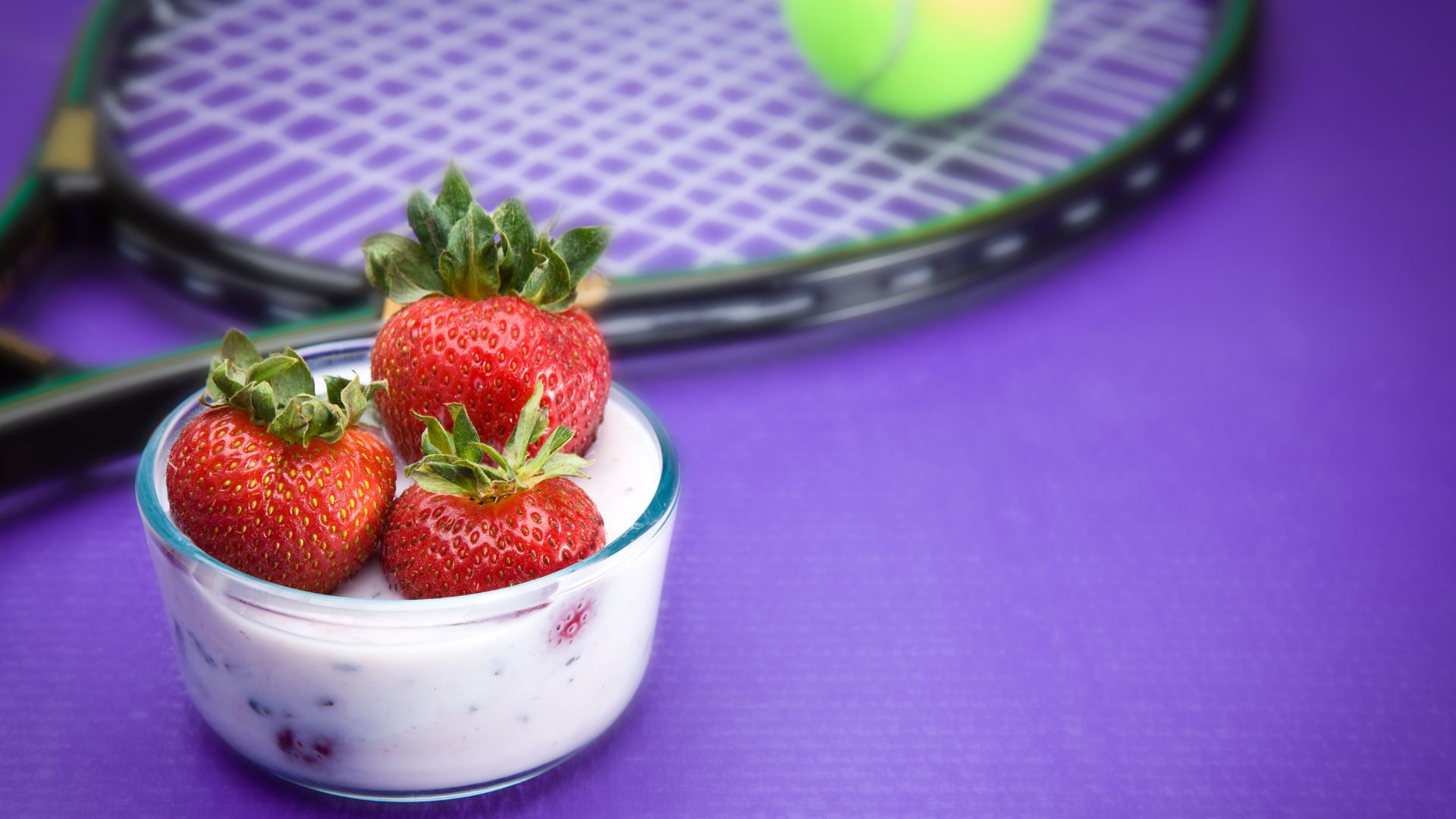 A bowl of fresh strawberries and cream sits on a purple surface next to a tennis racket and ball, suggesting a connection to the Wimbledon tennis tournament.