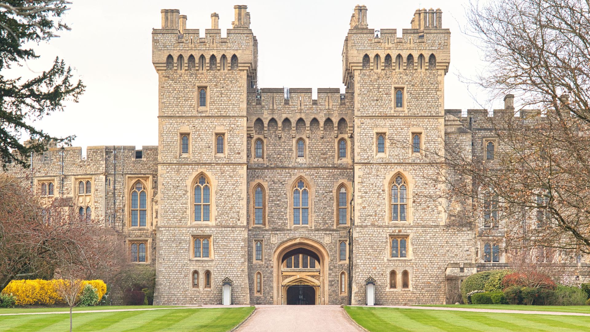 A grand stone castle with two prominent towers and numerous windows, set amidst a manicured green lawn and surrounded by trees, under a bright sky.