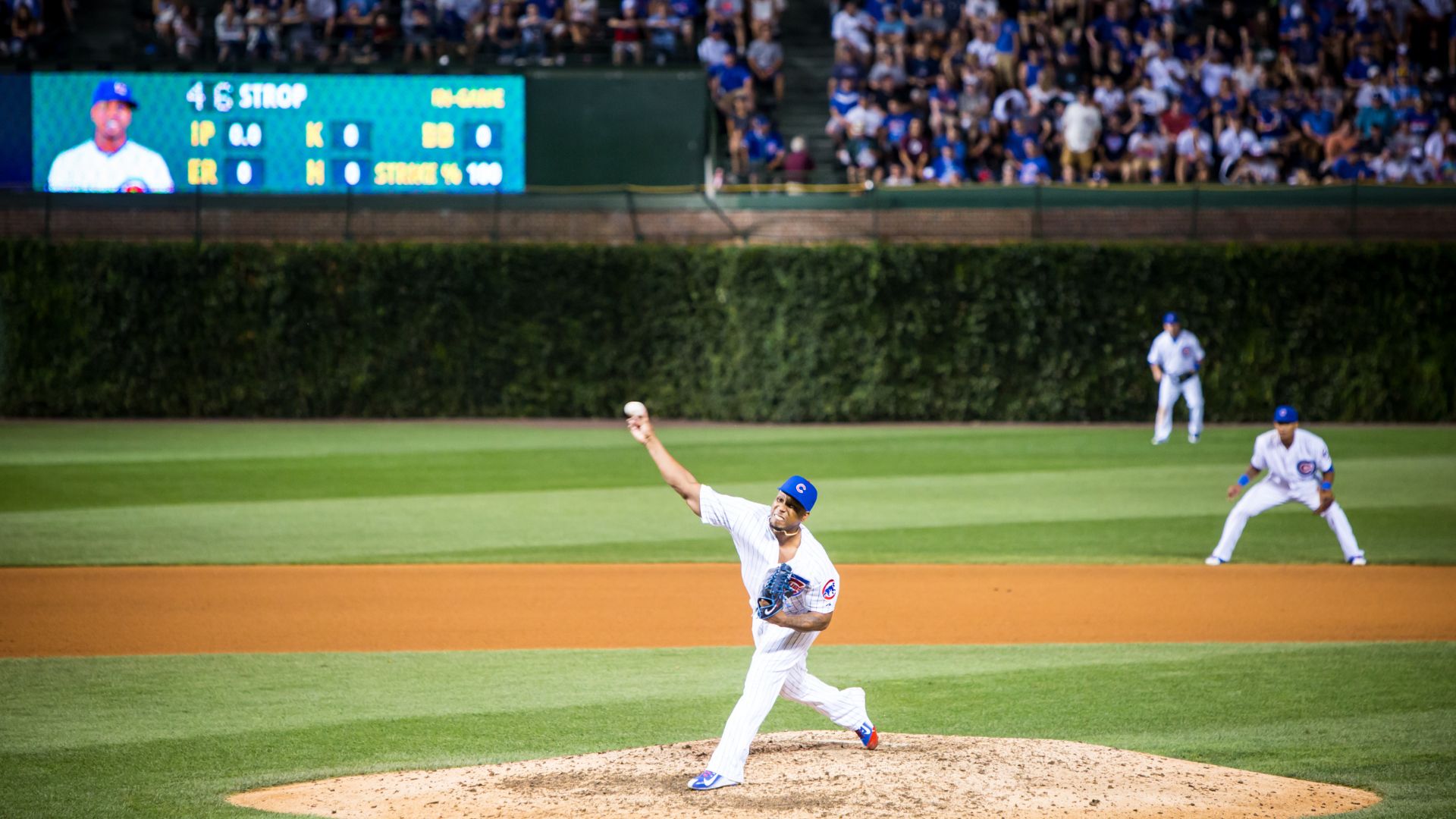 A wide shot of a baseball pitcher on the mound at Wrigley Field, mid-throw, with other players on the field and a scoreboard displaying "46 STROP" in the background, under stadium lights at night, with spectators in the stands. 
