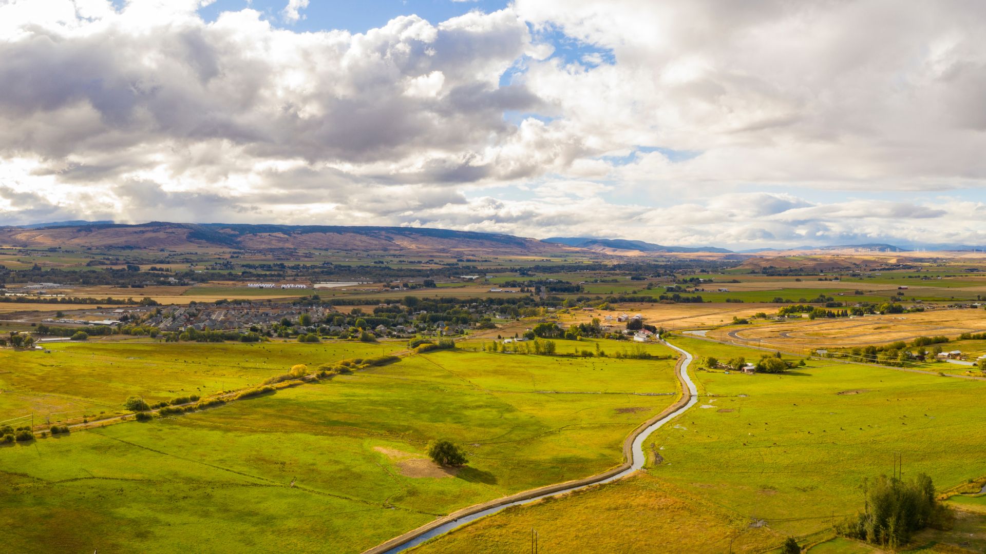 An aerial view of a verdant valley featuring a winding river or canal, surrounded by agricultural fields and distant hills under a cloudy sky.