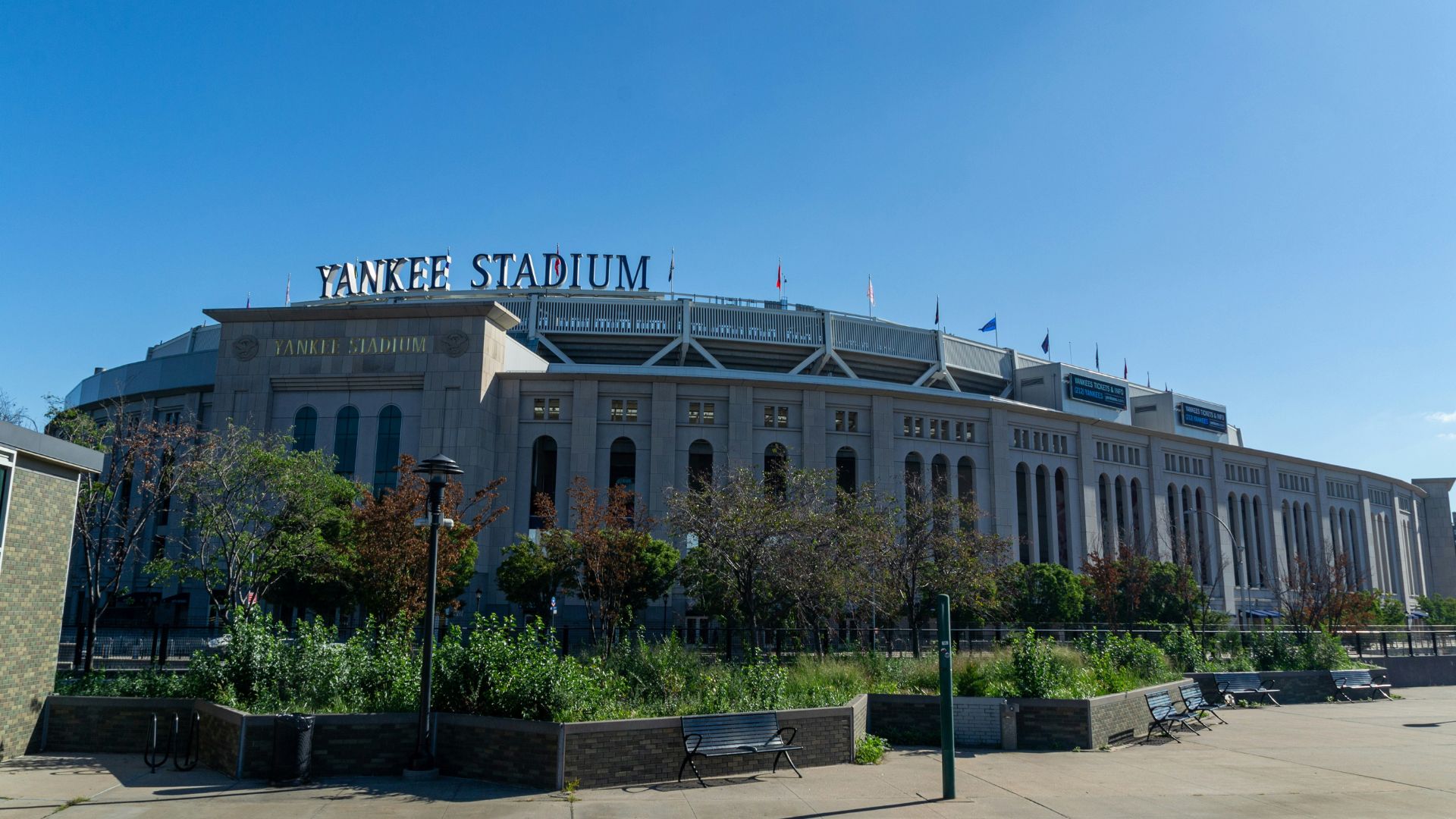 A wide exterior shot of Yankee Stadium on a clear day, showing its grand facade, surrounding green spaces, and a few benches in the foreground. 