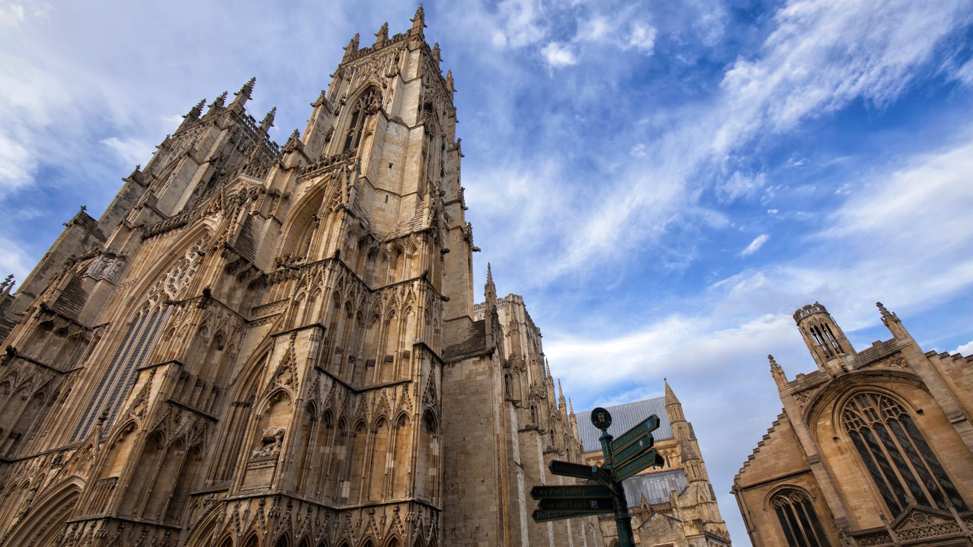 A low-angle, wide shot of the grand, Gothic-style York Minster cathedral under a bright sky with scattered clouds, featuring its intricate facade and towering spires, with a street signpost in the foreground and another building partially visible to the right.