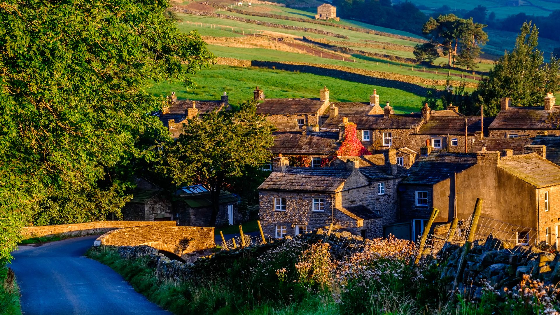 A picturesque stone village nestled in a green valley, with traditional stone houses, drystone walls, and a winding road leading towards a bridge, all set against a backdrop of rolling hills under warm, golden light.