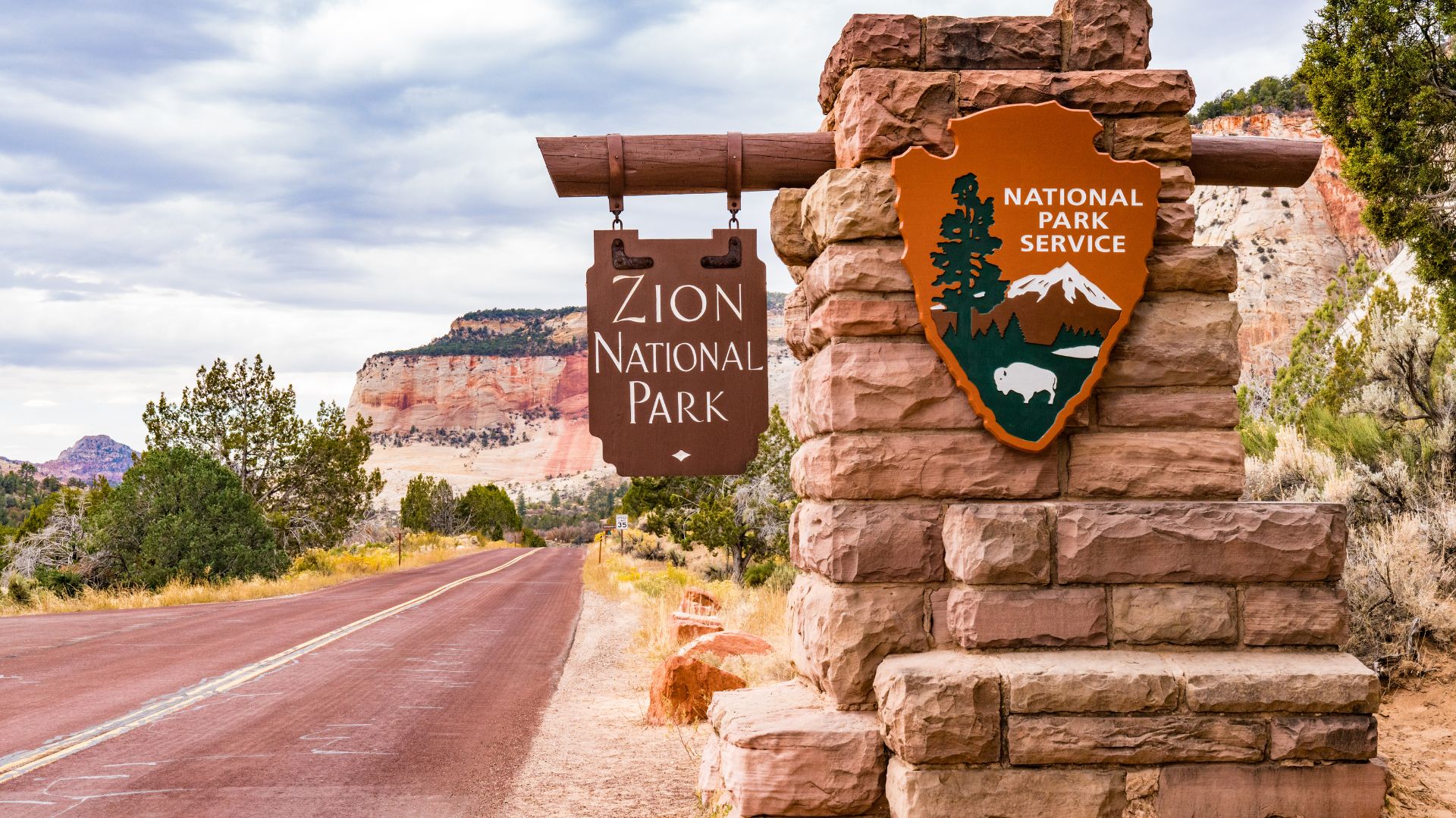 A roadside sign for Zion National Park, made of stone and wood, stands beside a paved road leading into a scenic canyon with towering sandstone cliffs under a cloudy sky. The sign features the National Park Service emblem and a separate wooden sign displaying "Zion National Park." 
