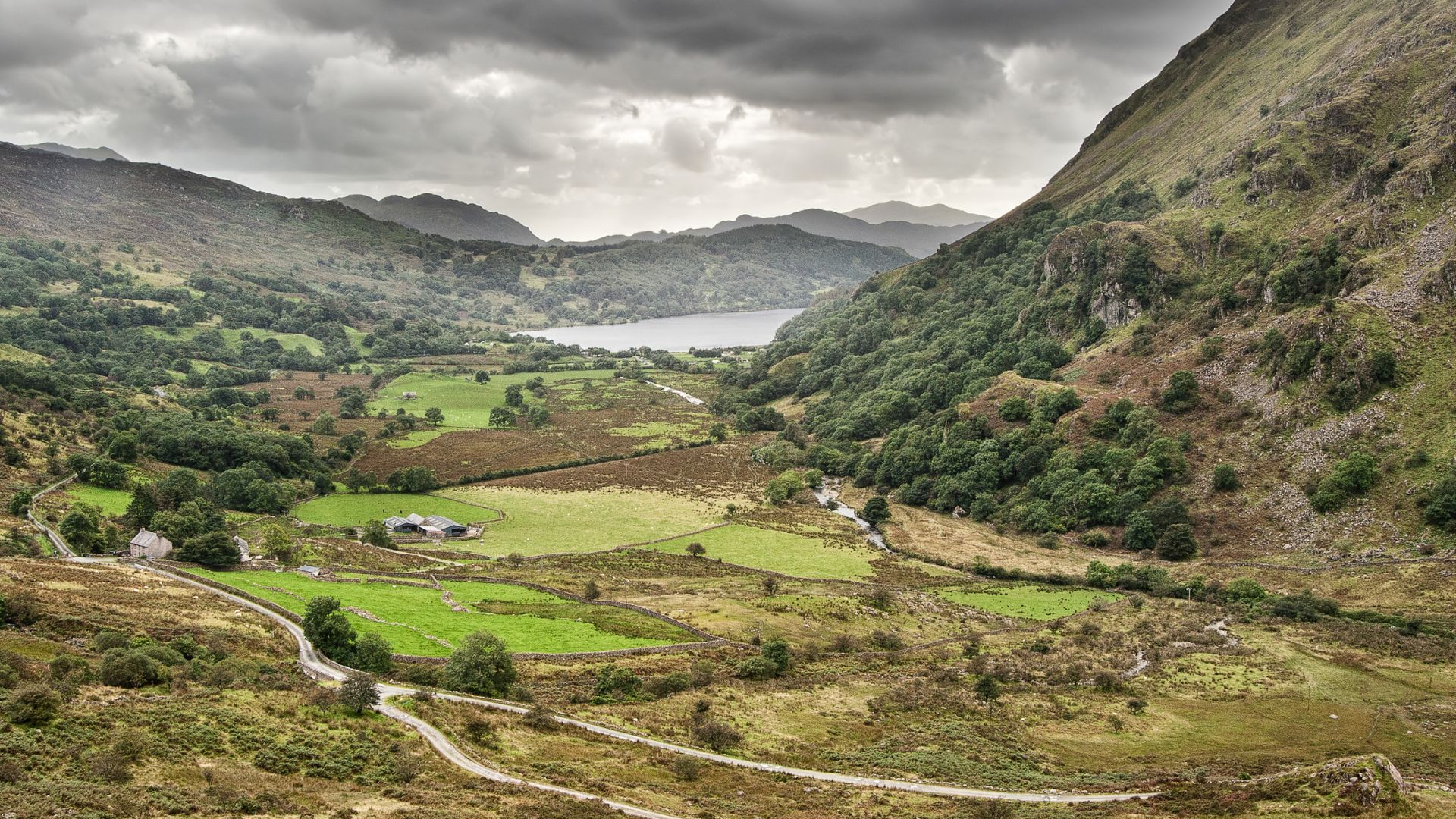 A panoramic view of a lush green valley, featuring a winding road, scattered fields, and a serene lake (Llyn Gwynant) nestled between verdant, rolling hills and towering mountains under a cloudy sky in Snowdonia National Park, Wales.