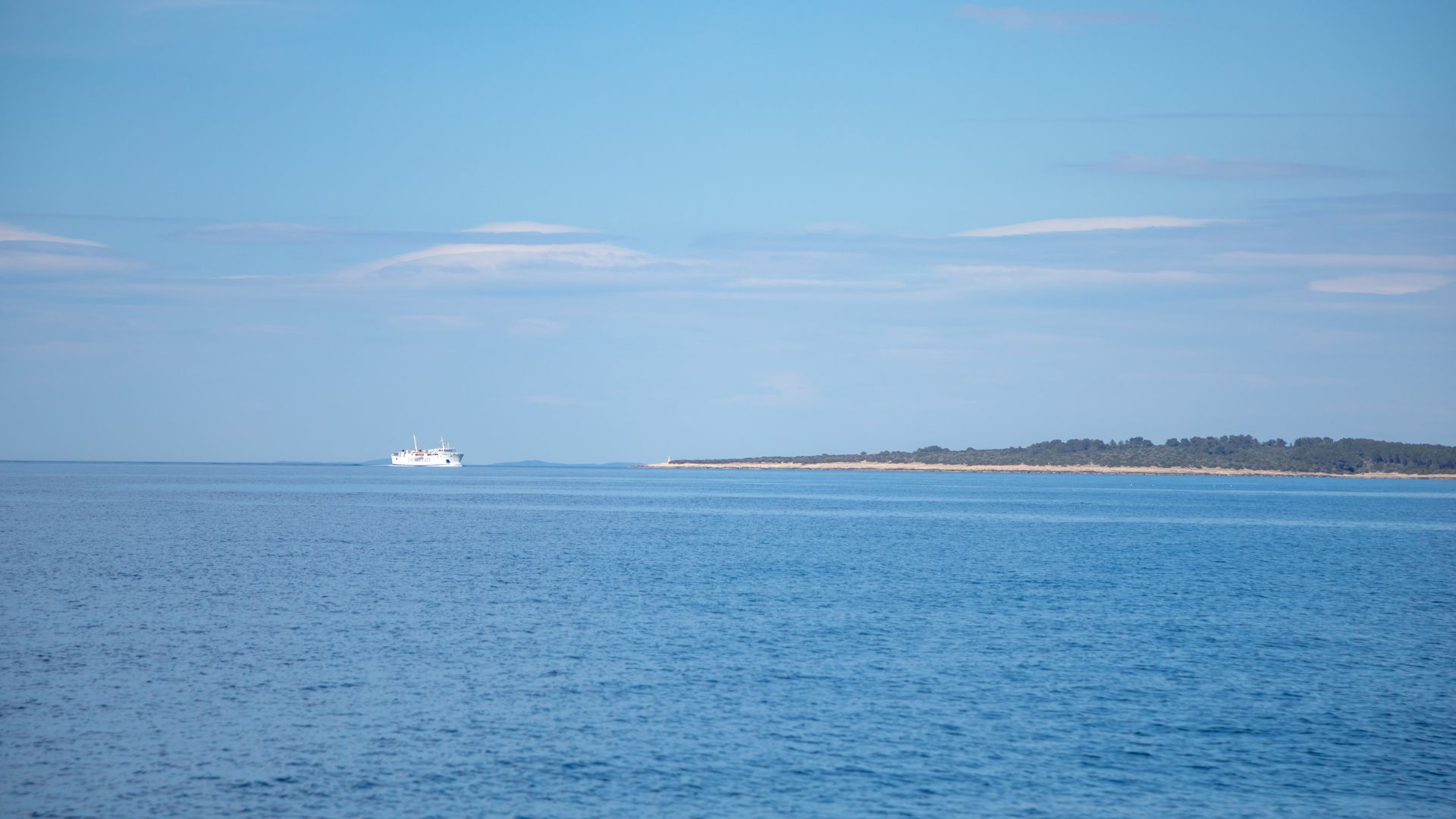 A white ferry boat sails across a calm blue sea under a clear sky, with a low-lying, forested landmass visible in the distance on the right.