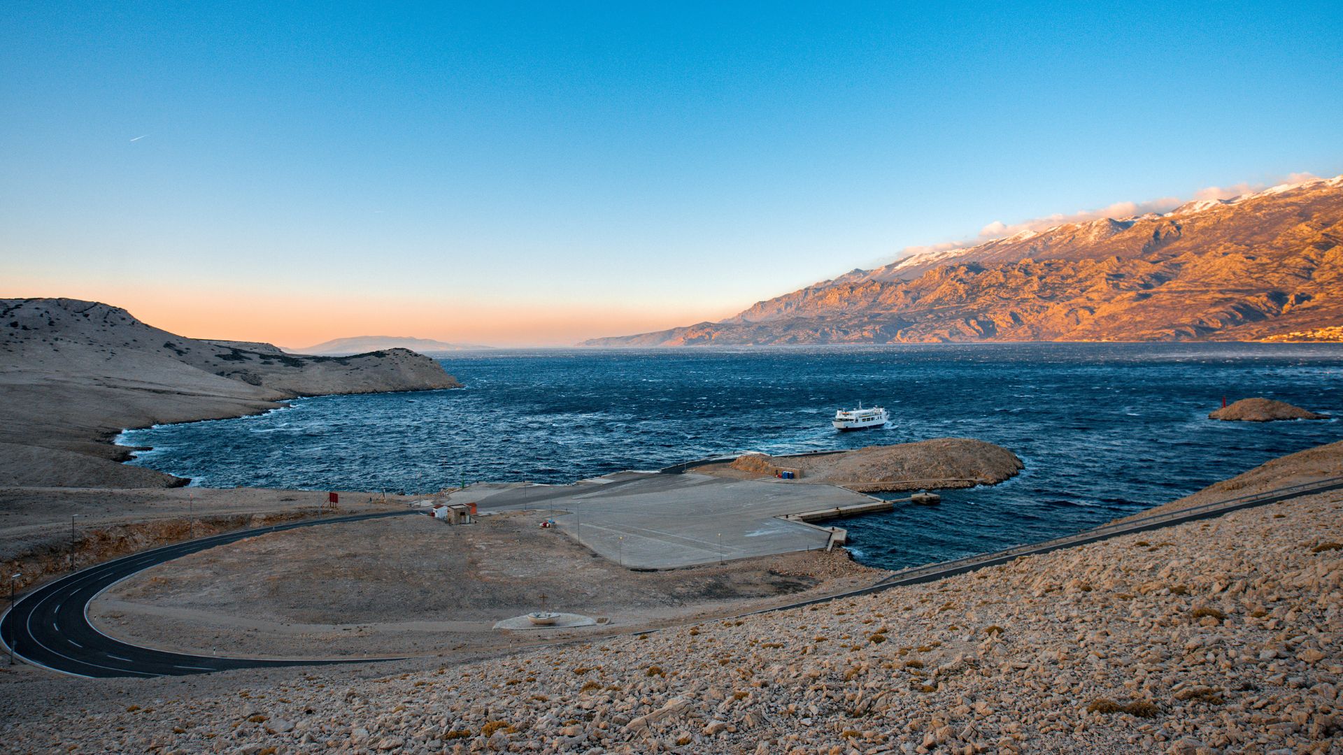 An aerial view of the rugged, moon-like landscape of Pag Island, Croatia, with a car ferry crossing the deep blue waters of the Adriatic Sea and mountains in the distance under a clear sky at sunset.