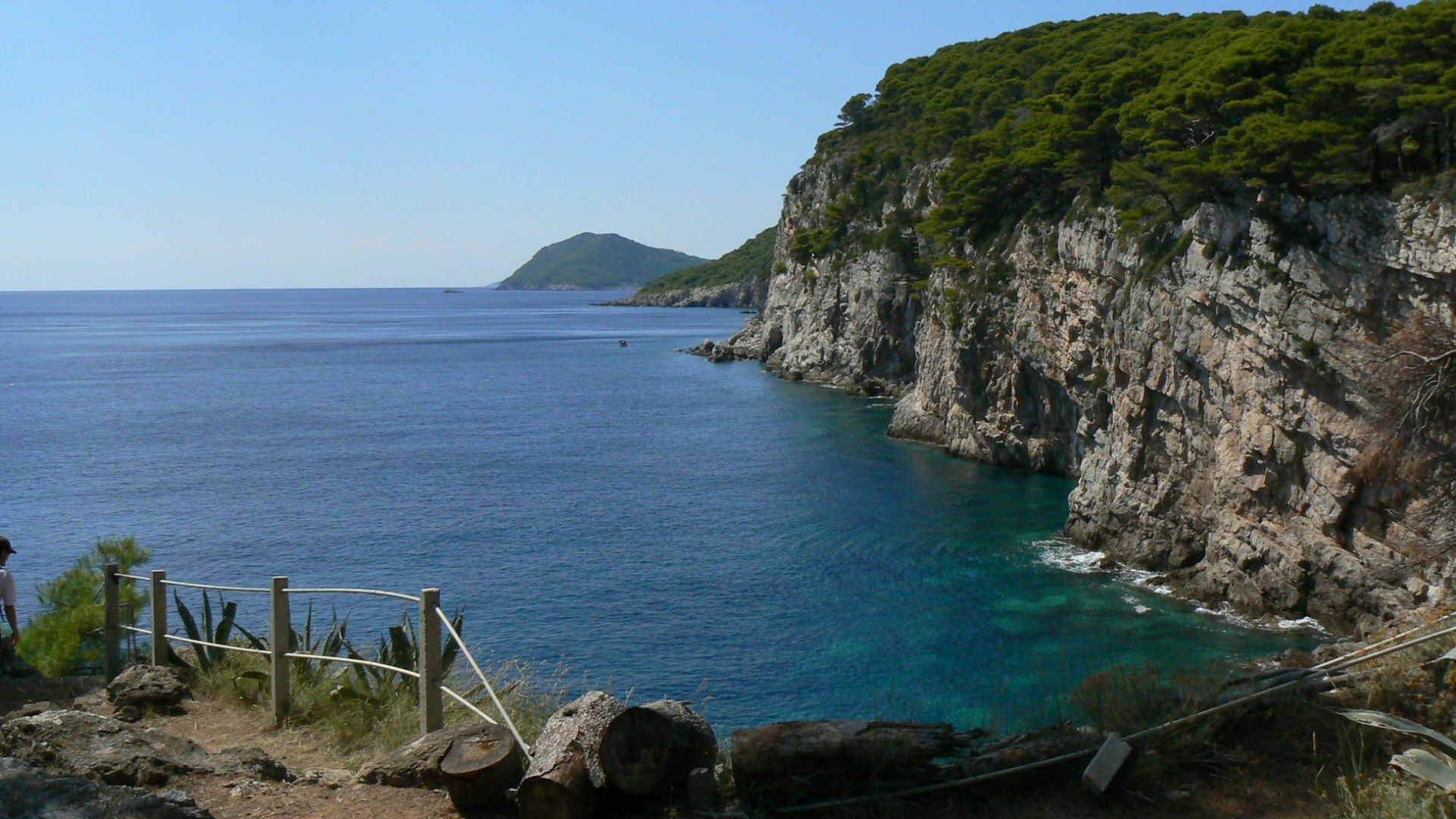 A vibrant blue sea with clear turquoise waters meets a rugged, tree-covered cliff face on the right, while a distant landmass is visible on the horizon. In the foreground, a rocky path with a simple rope fence offers a viewpoint overlooking the serene coastal scene.