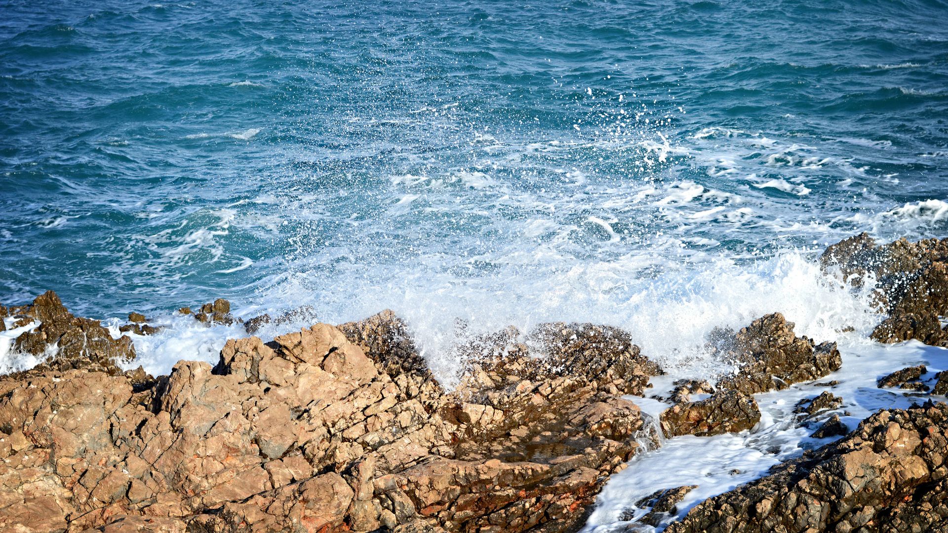 A high-angle view of vibrant blue ocean waves forcefully crashing against rugged, brown and orange-hued rocks on a sunny day, creating white spray and foam.