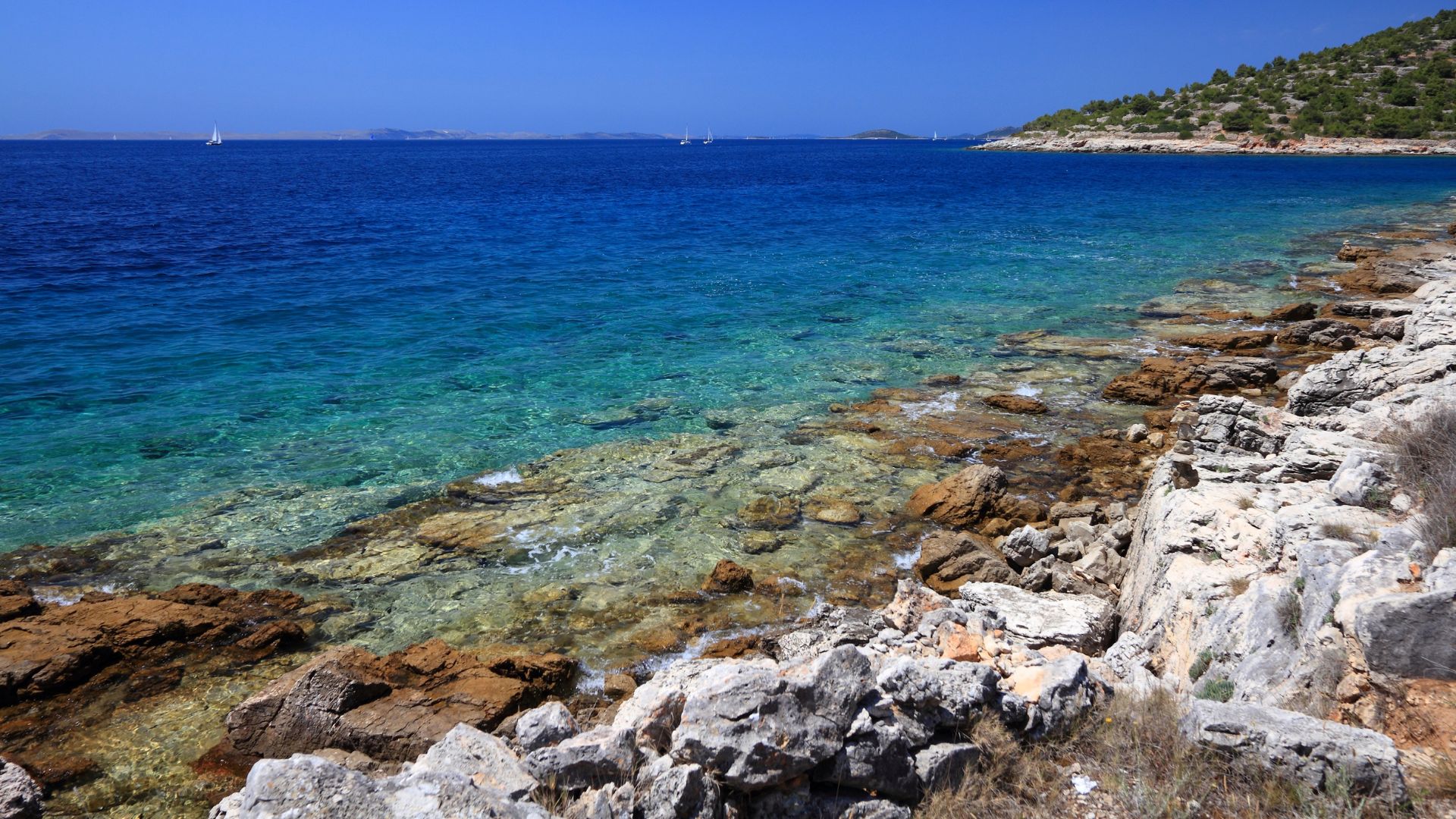 A wide-angle shot of a clear, turquoise sea meeting a rocky, rugged coastline under a bright blue sky. The water is shallow and transparent near the shore, revealing submerged rocks and a gradual transition to deeper, darker blue hues further out. Distant islands and a few boats are visible on the horizon.