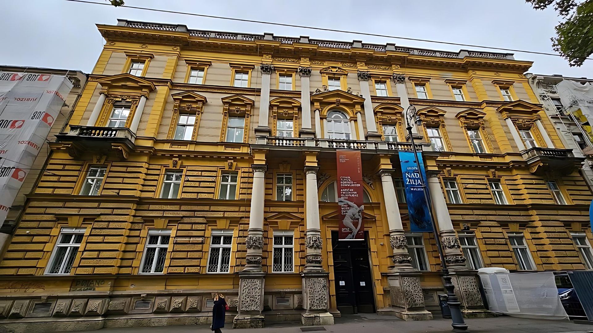 A grand, yellow and white neoclassical building, identified as the Archaeological Museum in Zagreb, Croatia, stands under an overcast sky with banners hanging from its facade and a person walking in front.