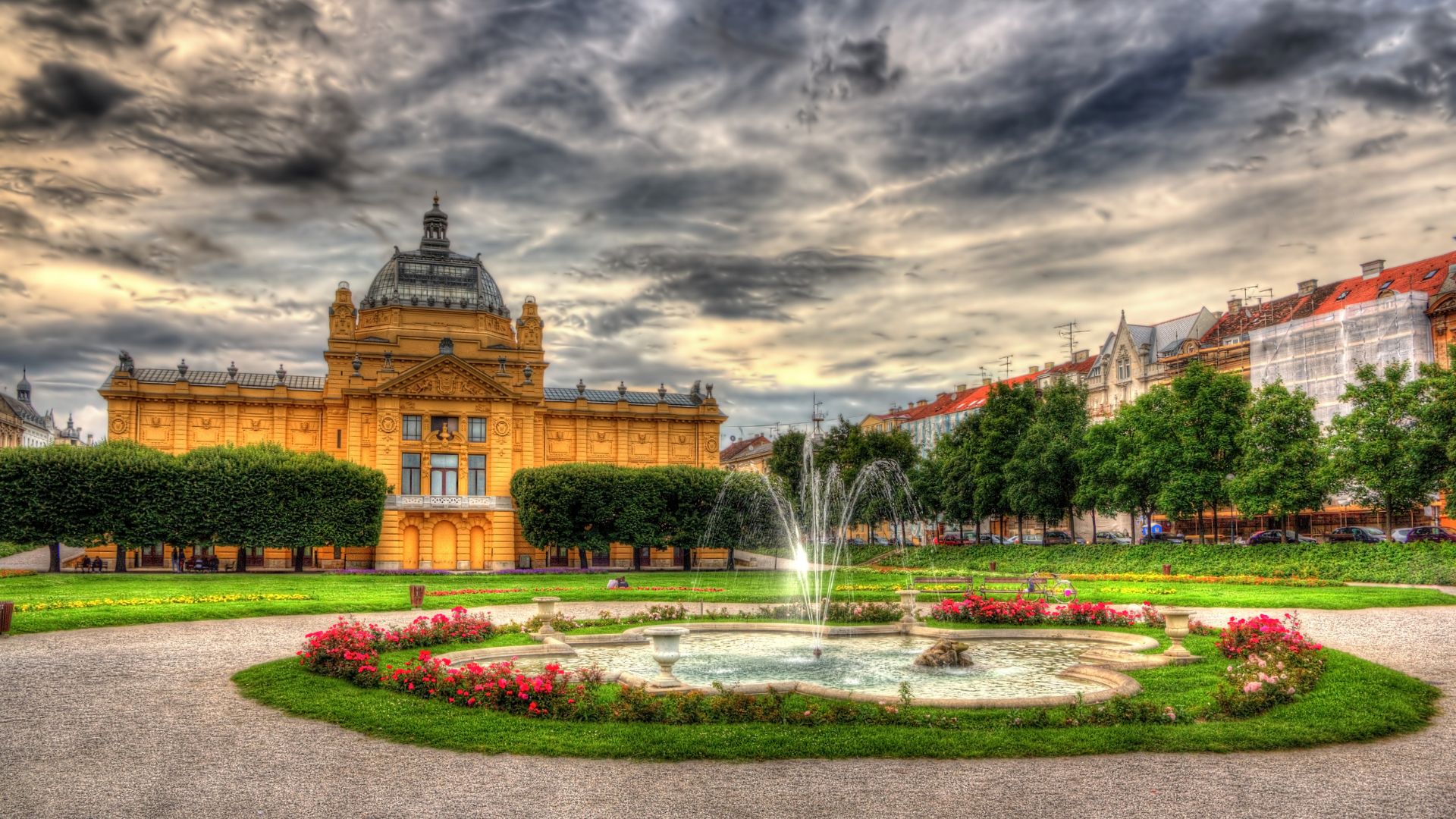 A vibrant, high-dynamic-range photograph capturing the yellow, domed Art Pavilion in Zagreb, Croatia, under a dramatic, cloudy sky. In the foreground, a decorative fountain with blooming red and pink flowers is set within a circular green lawn, leading to a paved path. Lush green trees line the area around the pavilion, and other buildings are visible in the background on the right. 
