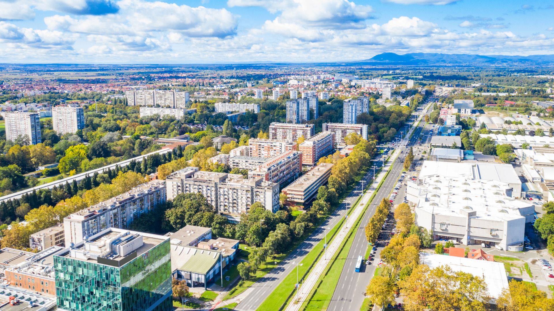 An aerial view of Avenue Dubrovnik in Novi Zagreb, Zagreb, Croatia, showing a wide multi-lane road flanked by numerous residential and commercial buildings, interspersed with green spaces and trees, under a partly cloudy sky.