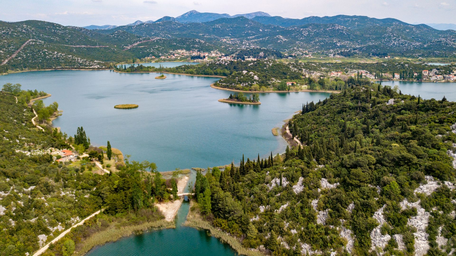 An aerial view of the Baćina Lakes in Croatia, showing a series of interconnected, clear blue lakes surrounded by lush green hills, dense forests, and a small town in the distance under a partly cloudy sky.