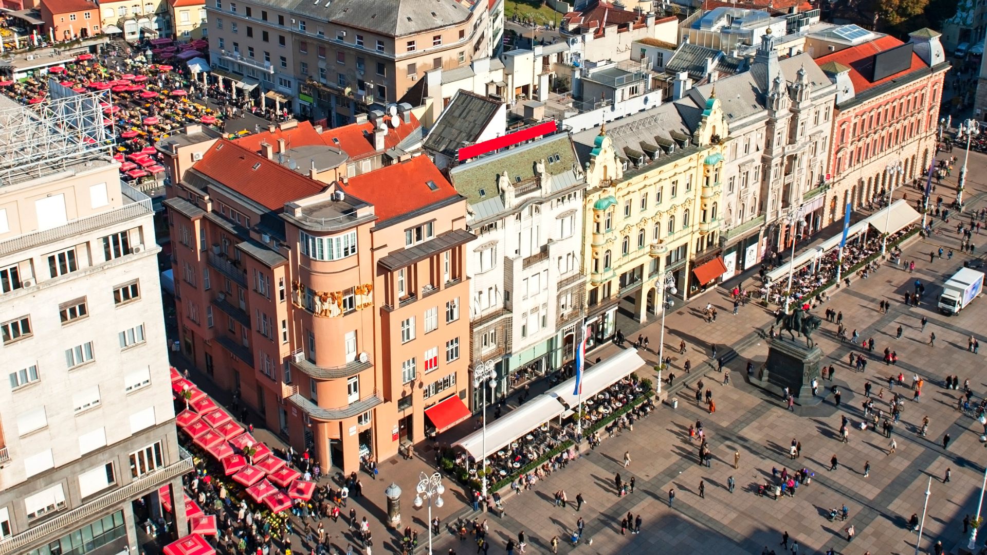 An aerial view of Ban Jelačić Square in Zagreb, Croatia, showing a bustling public square lined with colorful historic buildings, a statue, and a vibrant market area with red awnings. 
