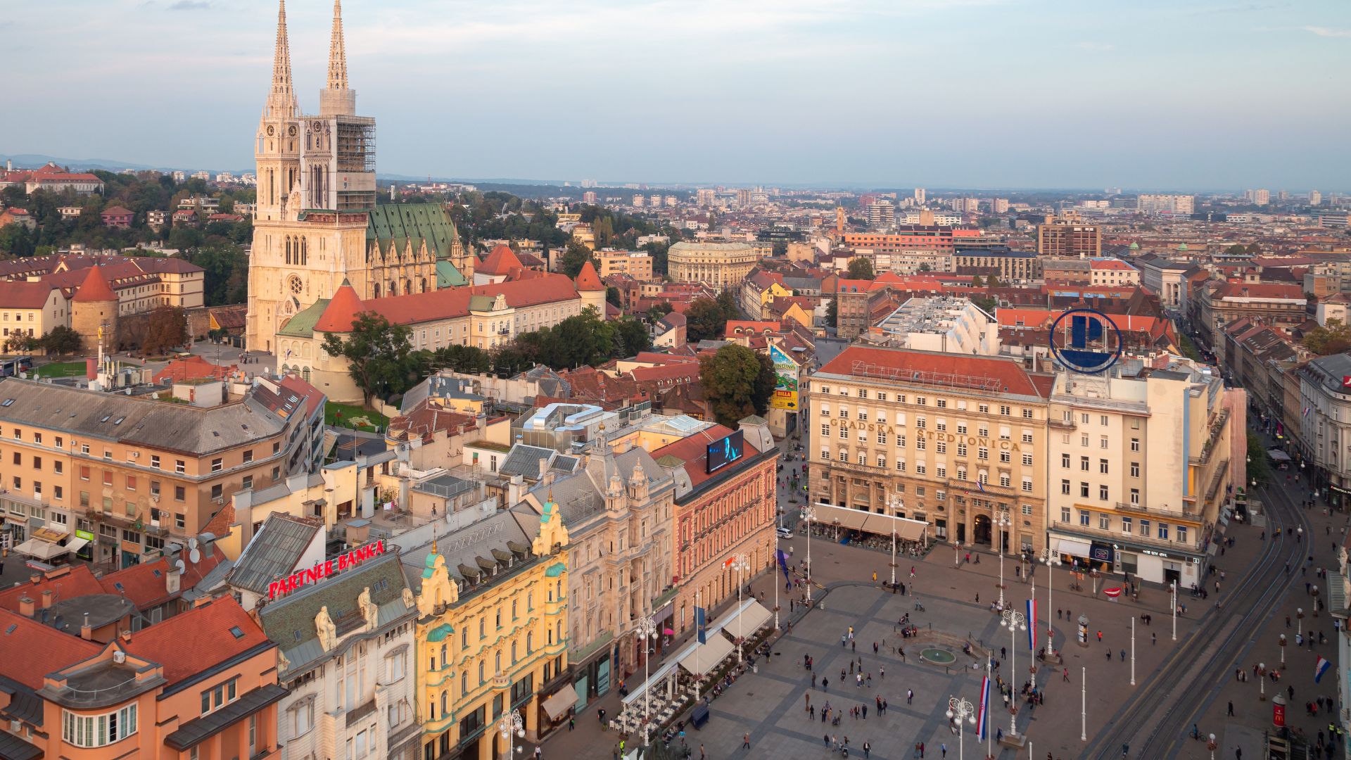 An aerial view of a bustling European city square, Ban Jelačić Square in Zagreb, Croatia, with historic buildings lining its edges, a large equestrian statue in the center, and a prominent cathedral with tall spires in the background.
