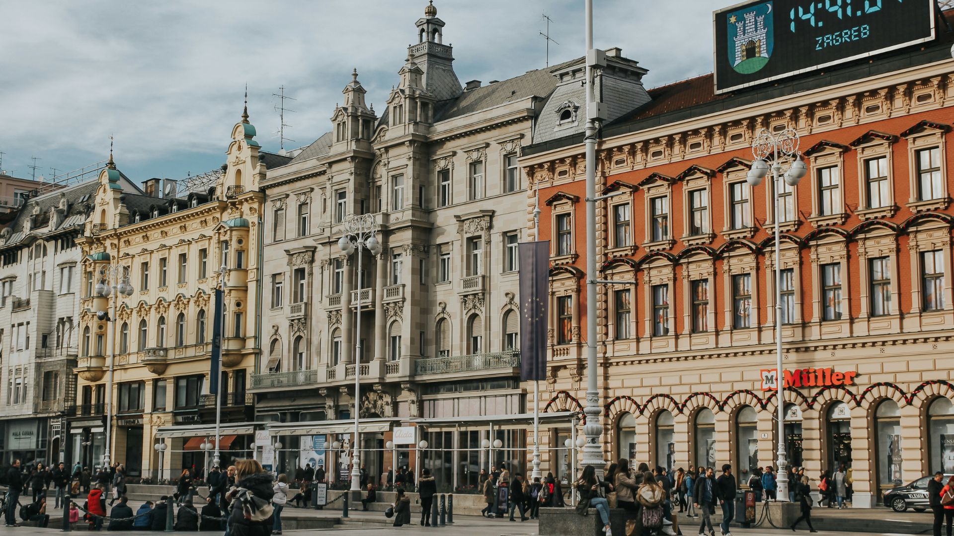 A wide-angle view of a bustling European city square, featuring historic buildings with detailed facades, numerous pedestrians, and a prominent digital clock displaying the time and "ZAGREB" against a cloudy sky.