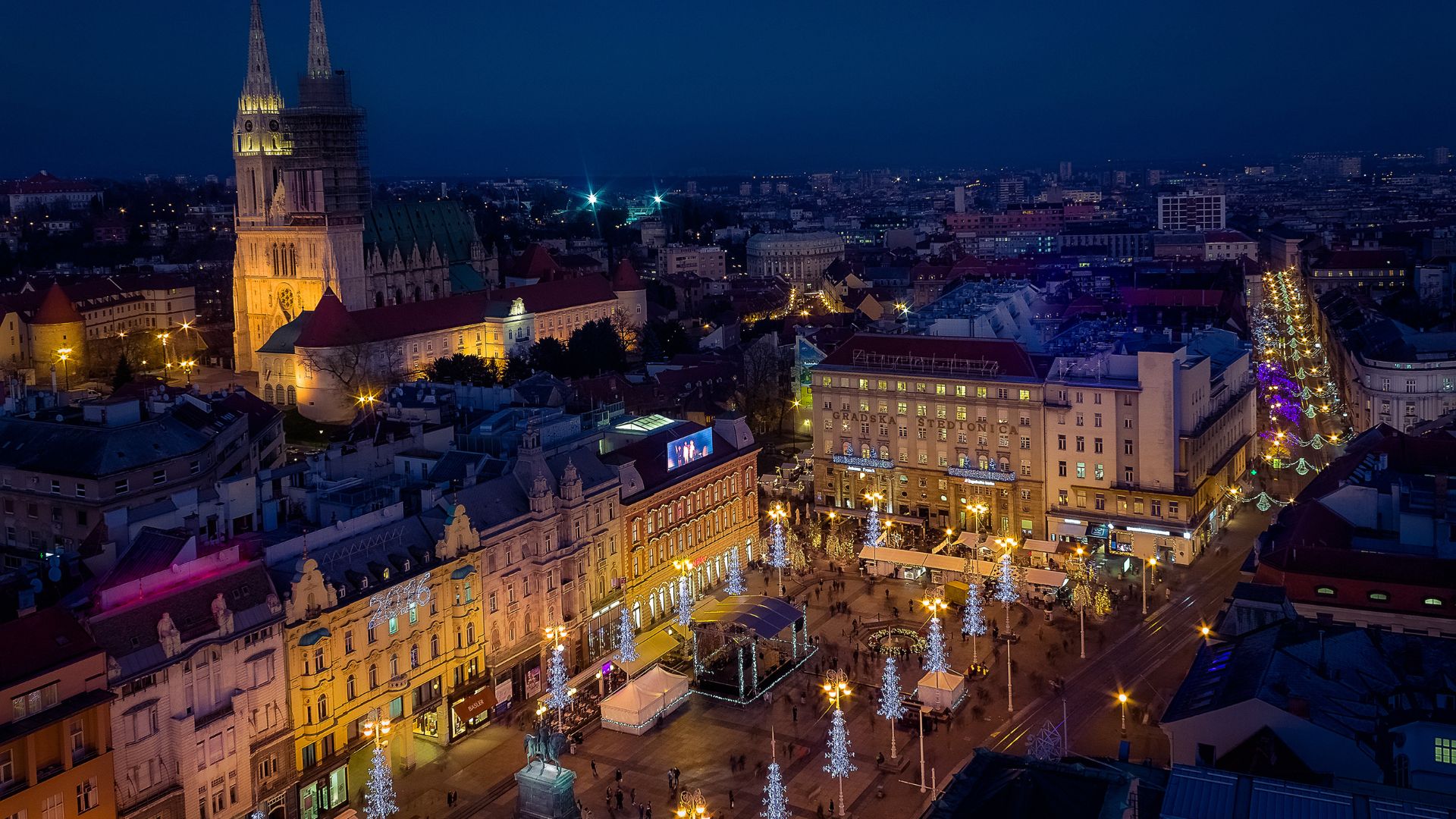 A nighttime aerial view of a bustling city square, Ban Jelačić Square in Zagreb, illuminated by streetlights and festive decorations, with historic buildings and a large cathedral visible in the background against a dark blue sky.