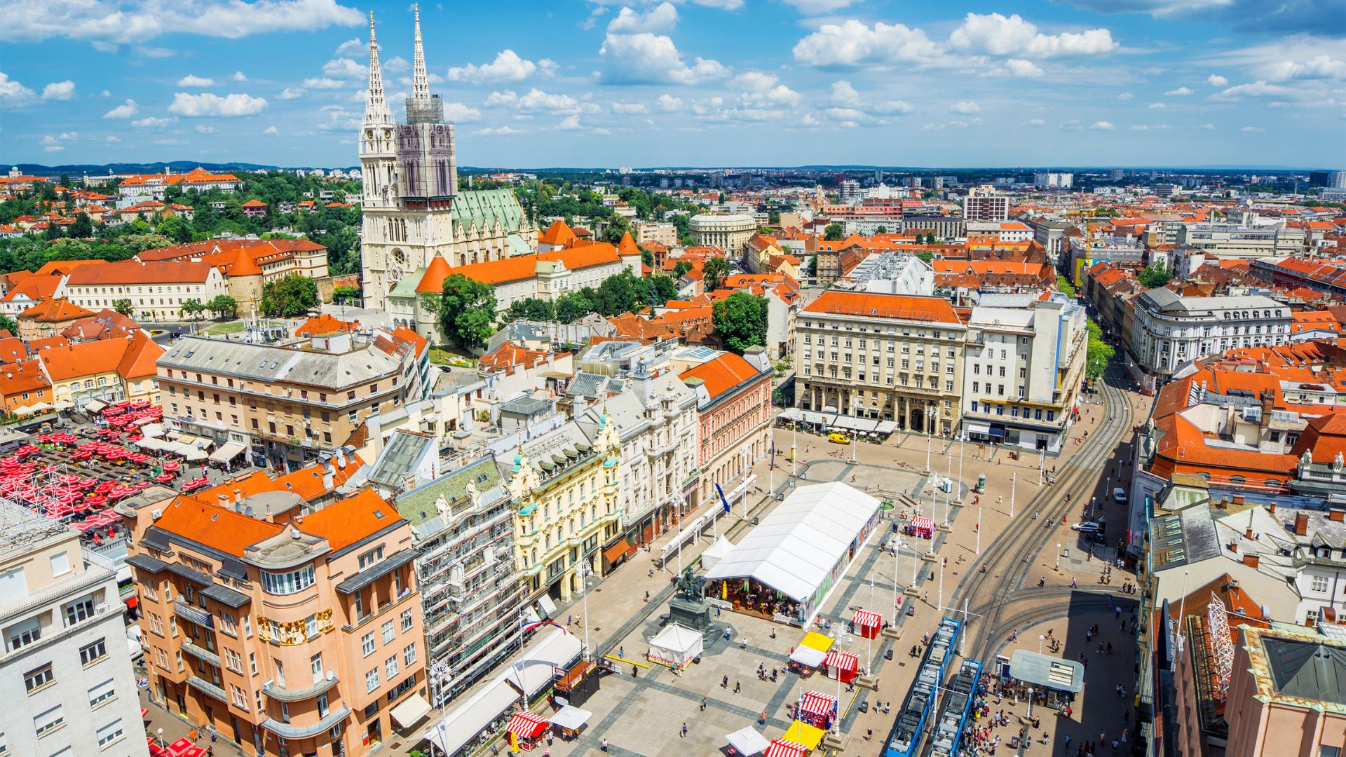An aerial view captures the bustling Ban Jelačić Square in Zagreb, Croatia, featuring a large pedestrian area with trams, surrounded by historic buildings with orange-tiled roofs, and the prominent Zagreb Cathedral visible in the background against a blue sky with scattered clouds.