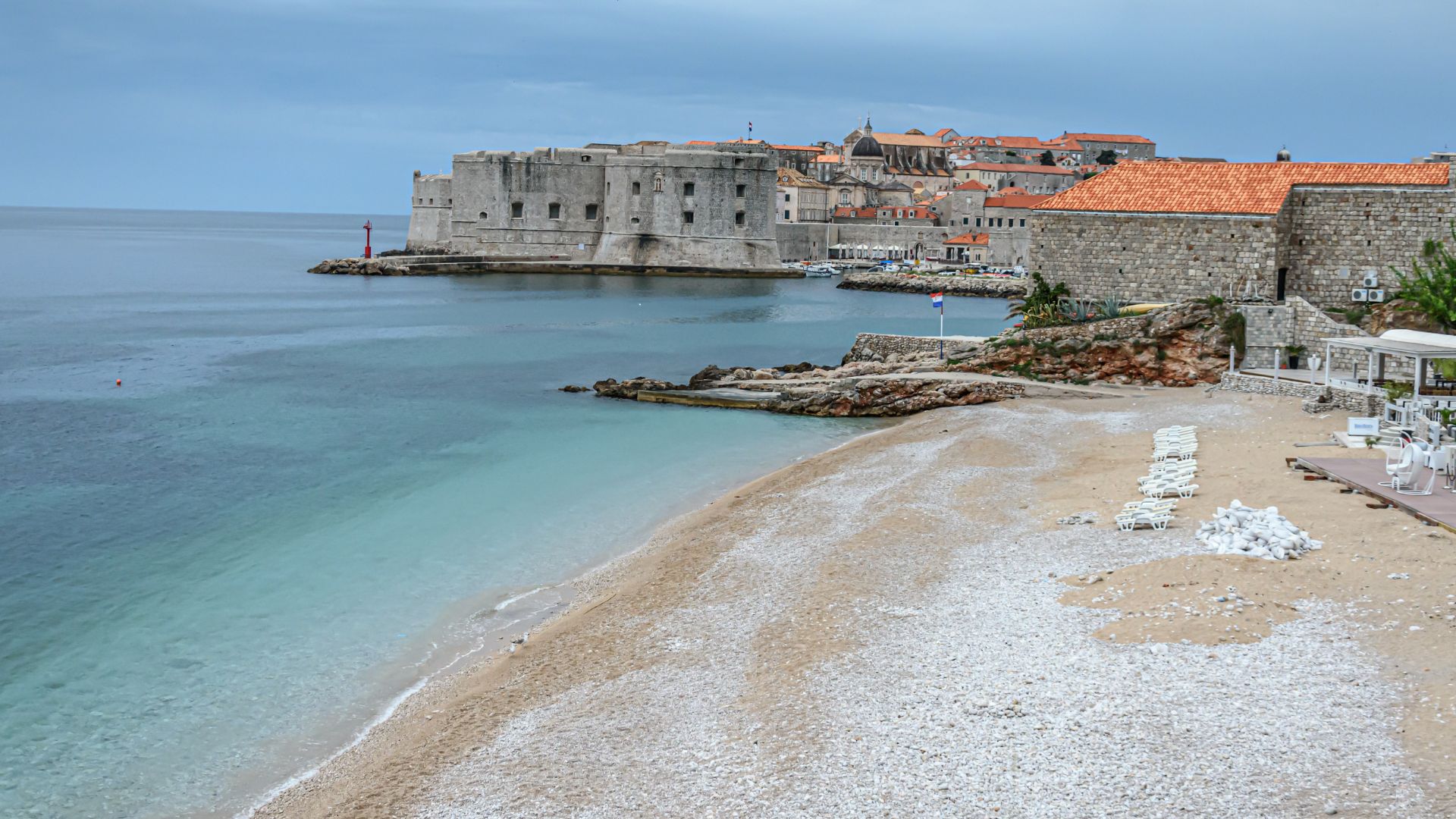 A panoramic view of Banje Beach in Dubrovnik, Croatia, with the clear turquoise waters of the Adriatic Sea in the foreground and the historic walled city of Dubrovnik in the background under a cloudy sky. The beach features light-colored pebbles and a few lounge chairs, while the city walls and buildings with red-tiled roofs are prominent on the coastline.