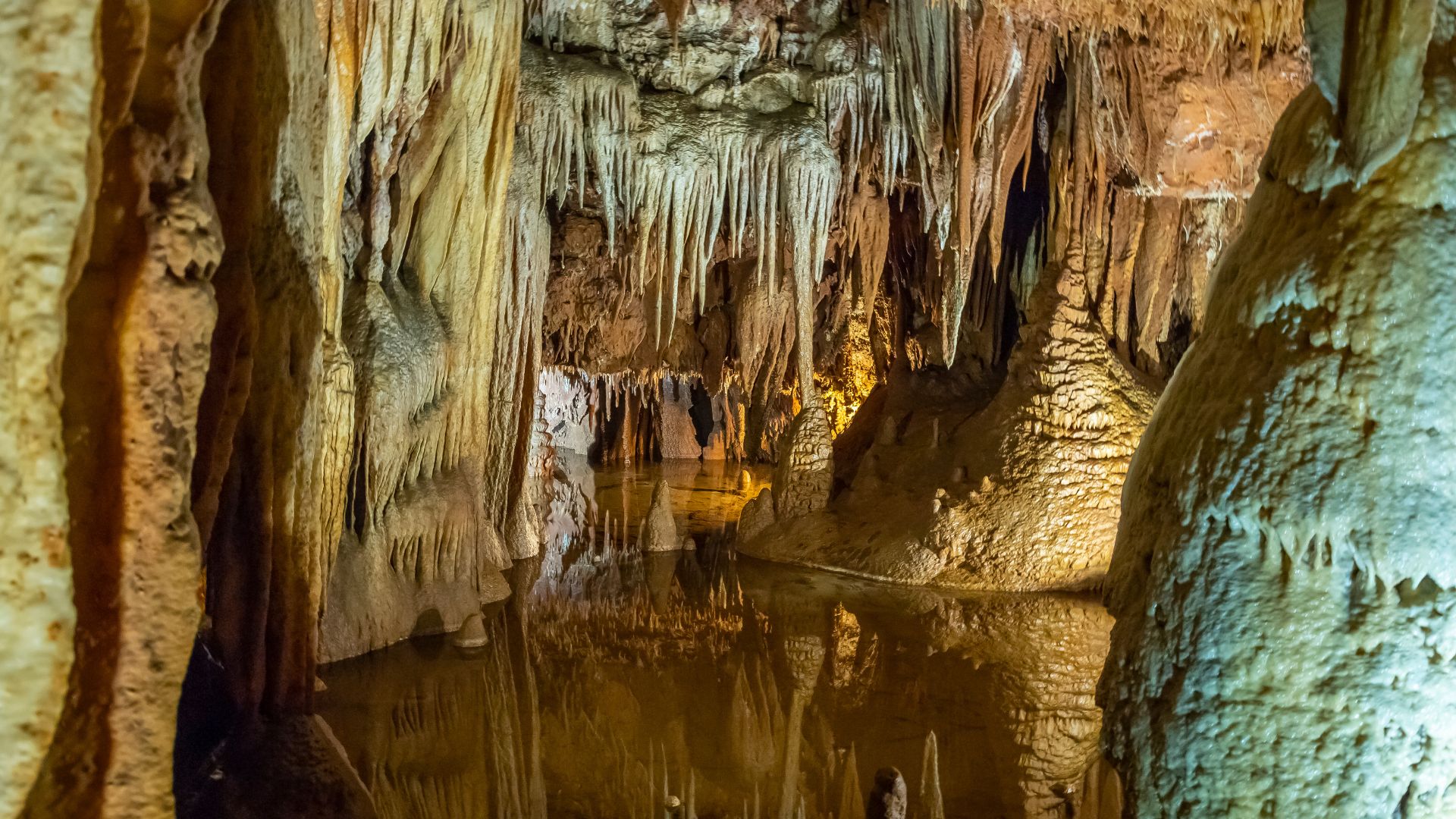 A dimly lit cave interior features numerous stalactites hanging from the ceiling and stalagmites rising from the floor, surrounding a calm, reflective underground lake. The formations display various earthy tones, and the water's surface mirrors the cave's natural architecture.