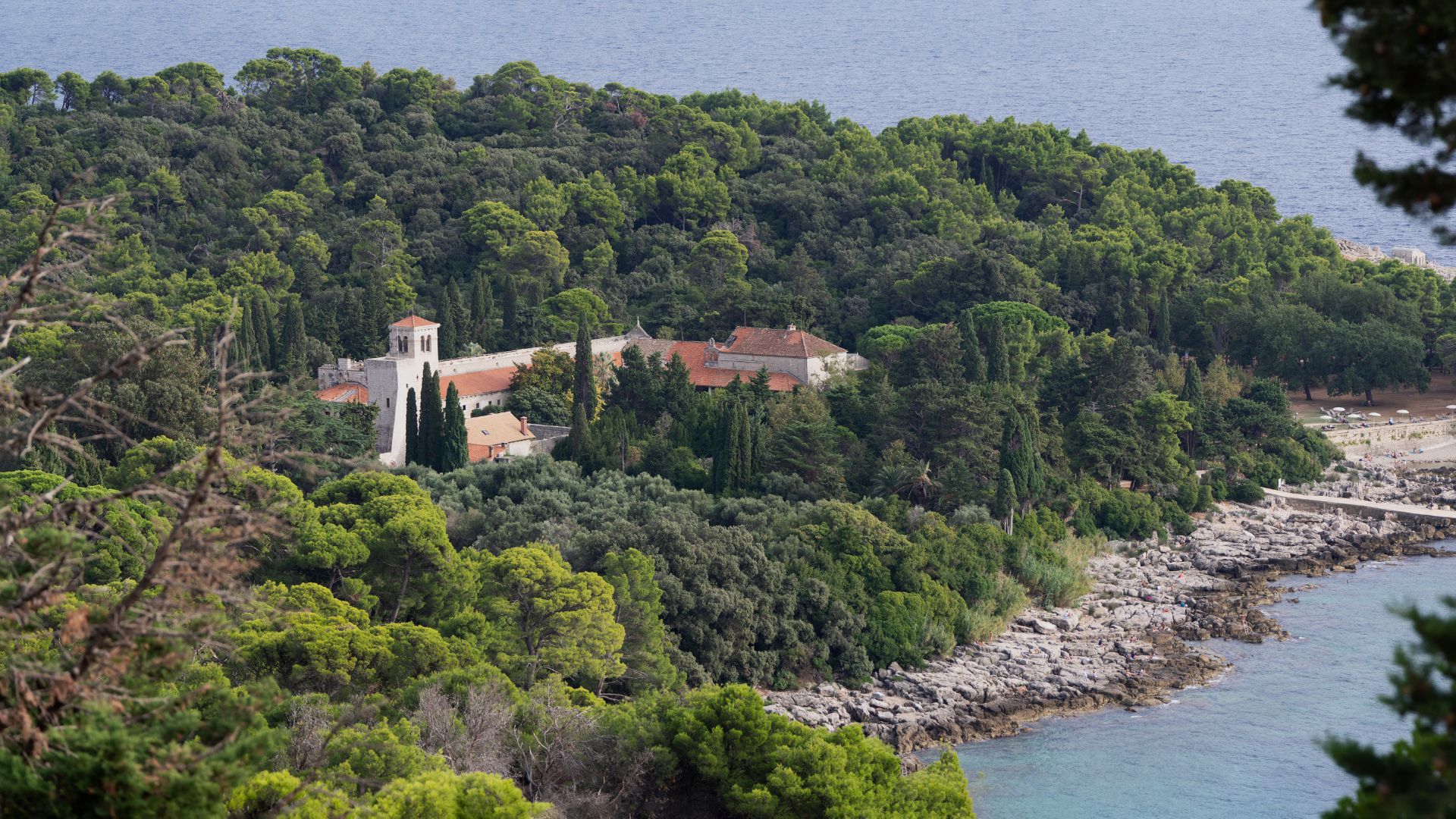 An aerial view of the Benedictine Monastery of St. Mary nestled amidst dense green foliage on Lokrum Island, with the rocky coastline and the Adriatic Sea visible in the foreground and background. 