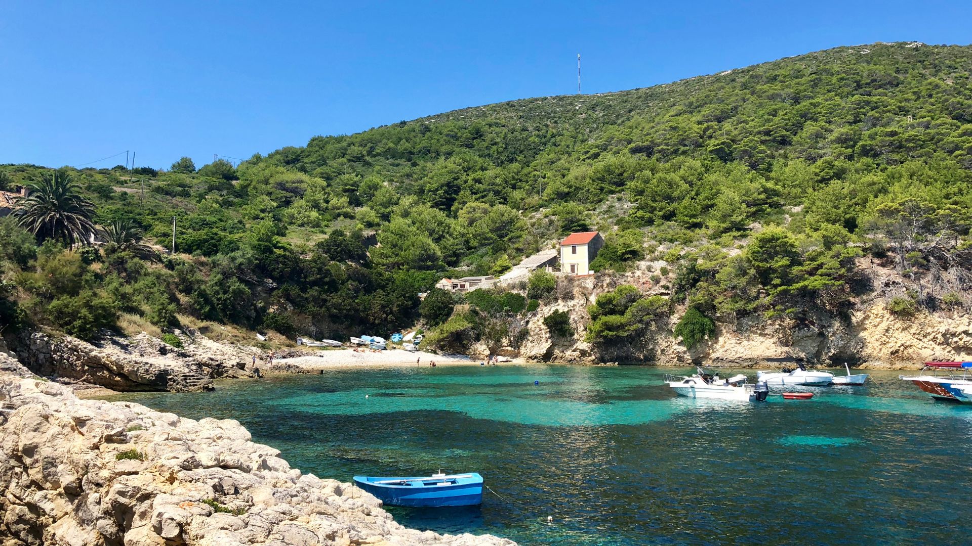 A picturesque cove with turquoise water, a small sandy beach, and several boats docked. A house sits atop a lush green, tree-covered cliff overlooking the bay under a clear blue sky.