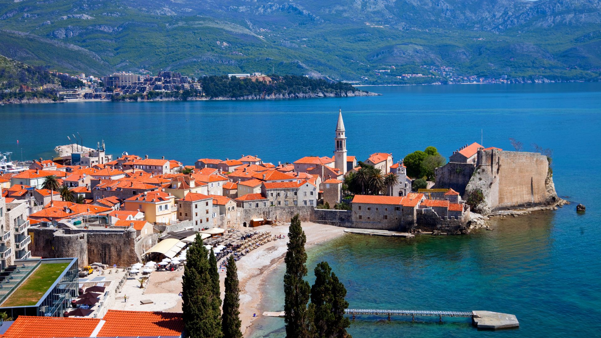 An aerial view of the historic Old Town of Budva, Montenegro, featuring red-tiled roofs, a prominent church spire, and ancient city walls, nestled along a clear blue bay with a sandy beach and lush green mountains in the background.