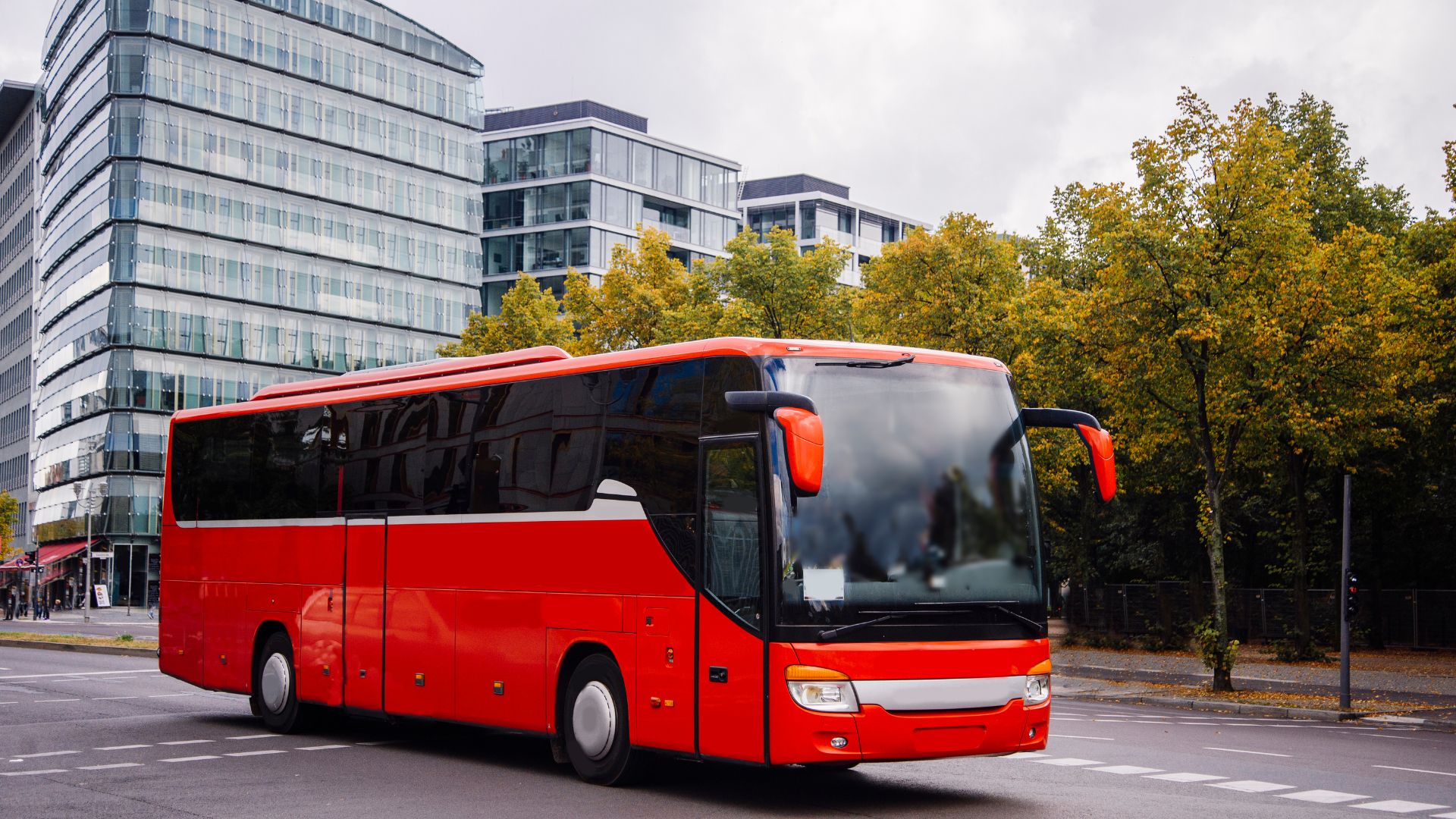 A bright red modern tour bus with dark tinted windows is parked on a city street, with contemporary buildings and green and autumnal trees in the background.
