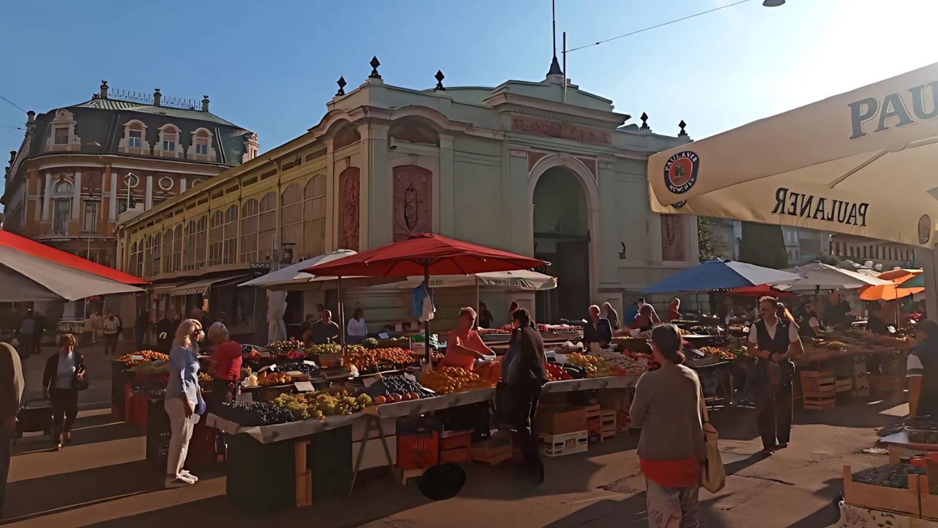 A vibrant outdoor market scene in front of an ornate, historic market hall building, with numerous stalls displaying fresh produce like fruits and vegetables, and people browsing the goods under a clear sky.