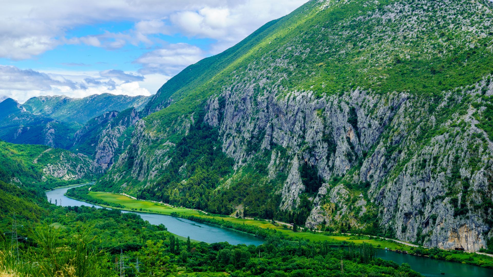 A vibrant landscape view of the Cetina River flowing through a deep, verdant canyon, flanked by towering, rocky cliffs and lush green mountains under a bright blue sky with scattered clouds.