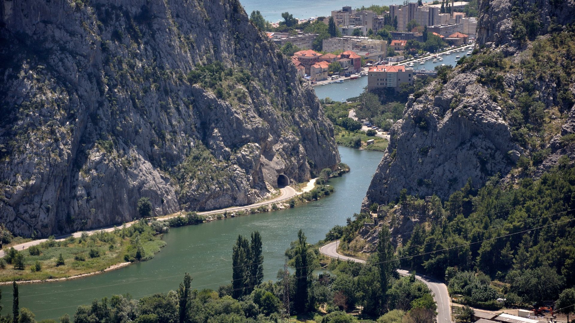 An aerial view shows the Cetina River winding through a deep, narrow canyon with steep, rocky cliffs covered in sparse vegetation on both sides. A road with a tunnel entrance runs along the riverbank on the left, and a winding road is visible on the right cliff face, leading towards a town in the distance at the river's mouth.