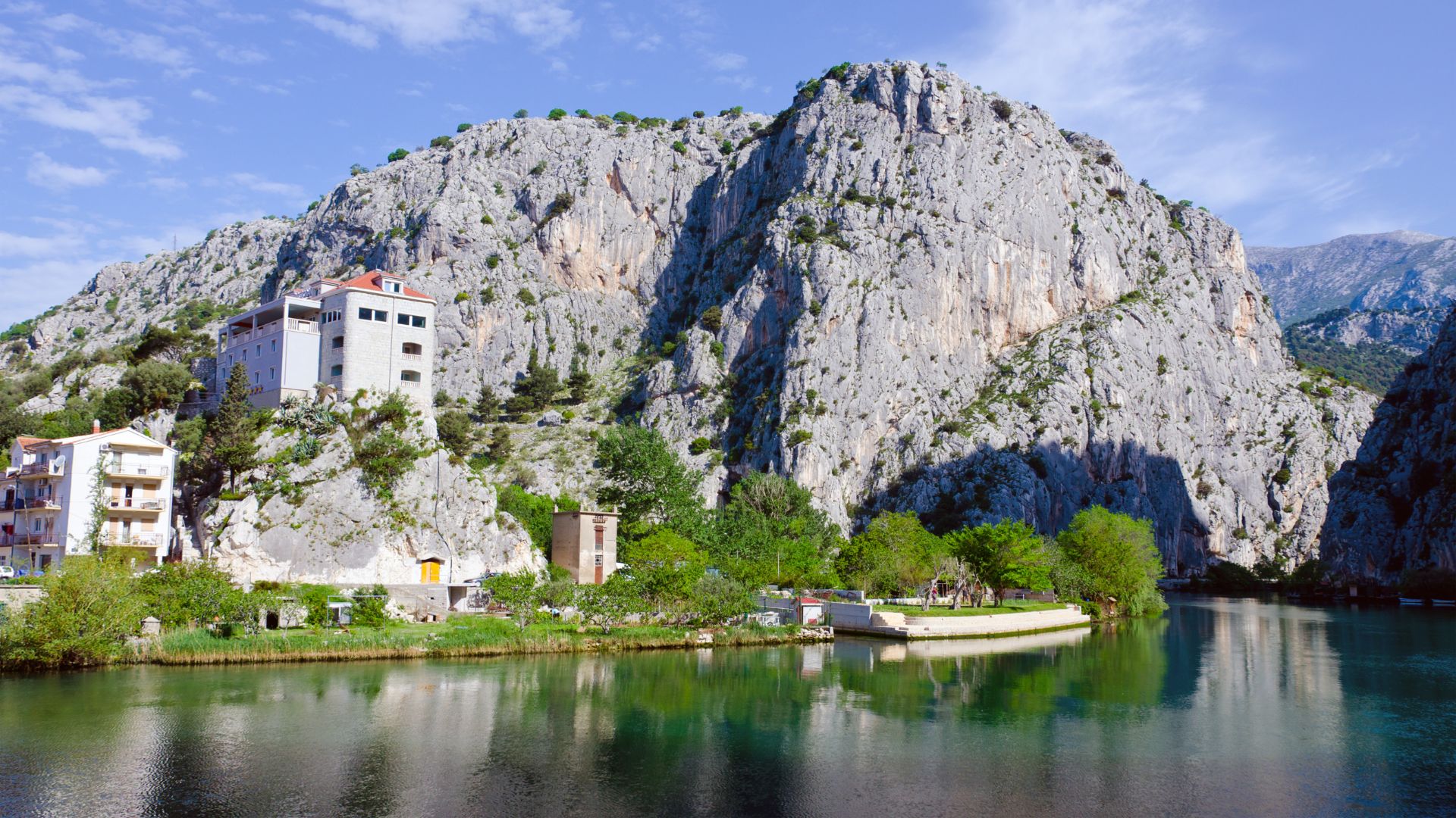 A picturesque view of the Cetina River in Omiš, Croatia, with a large rocky mountain face on the right, buildings including the Hotel Villa Dvor on the left bank, and lush greenery reflecting in the calm river waters under a bright blue sky.
