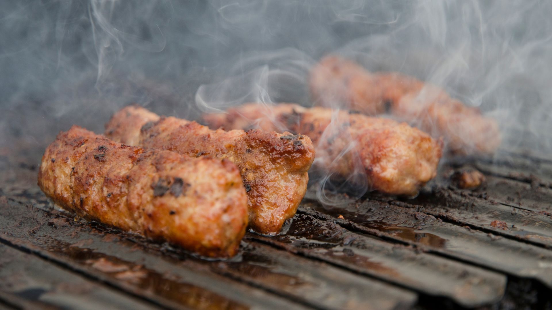 Close-up of several pieces of grilled, browned minced meat sausages (likely ćevapi or mici) cooking on a smoky barbecue grill.