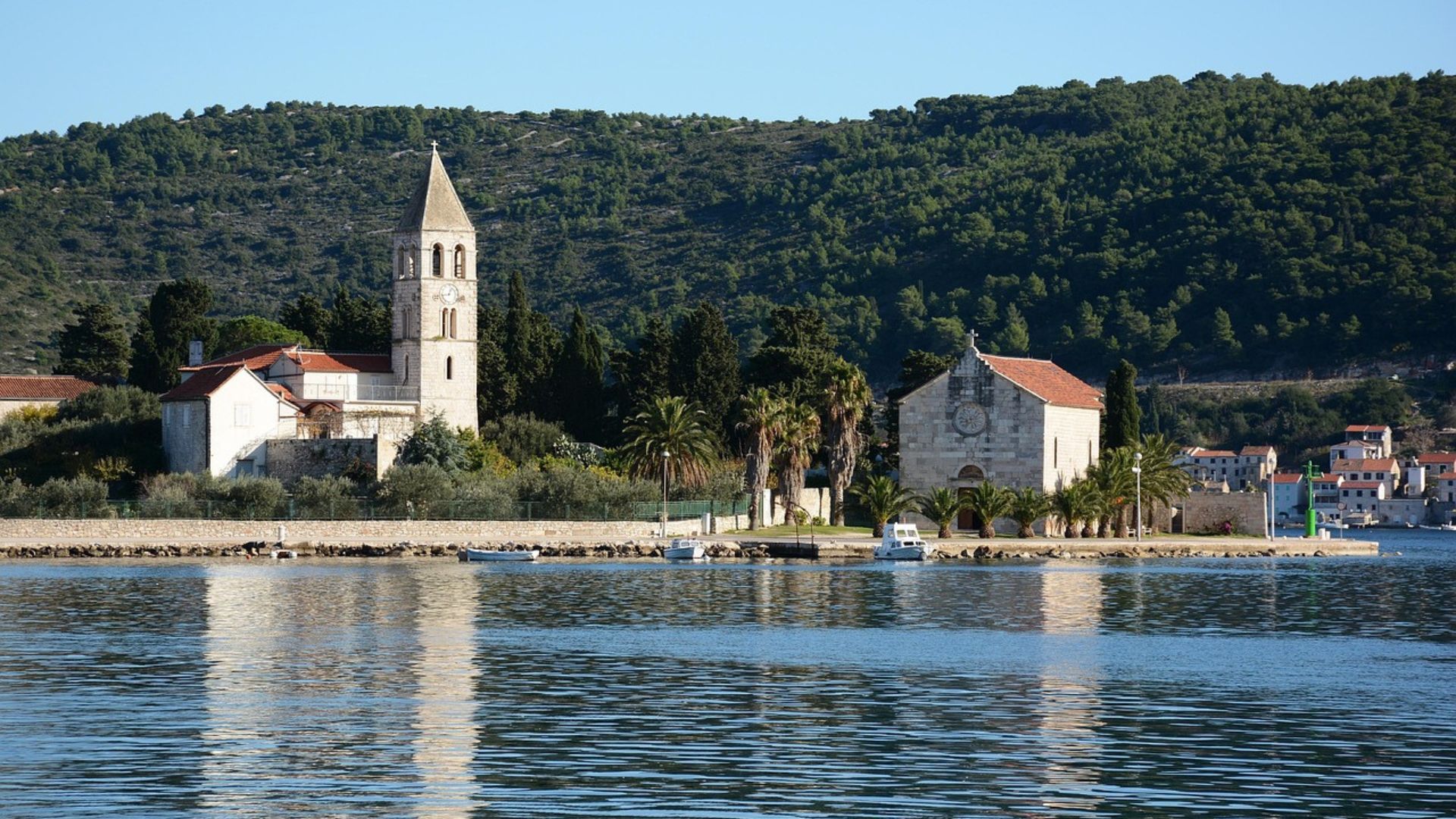 A serene coastal scene showing the Church of St. Jerome and Franciscan Monastery with its distinctive bell tower on the island of Vis, Croatia, reflected in the calm waters of the Adriatic Sea, with a lush green hillside in the background.