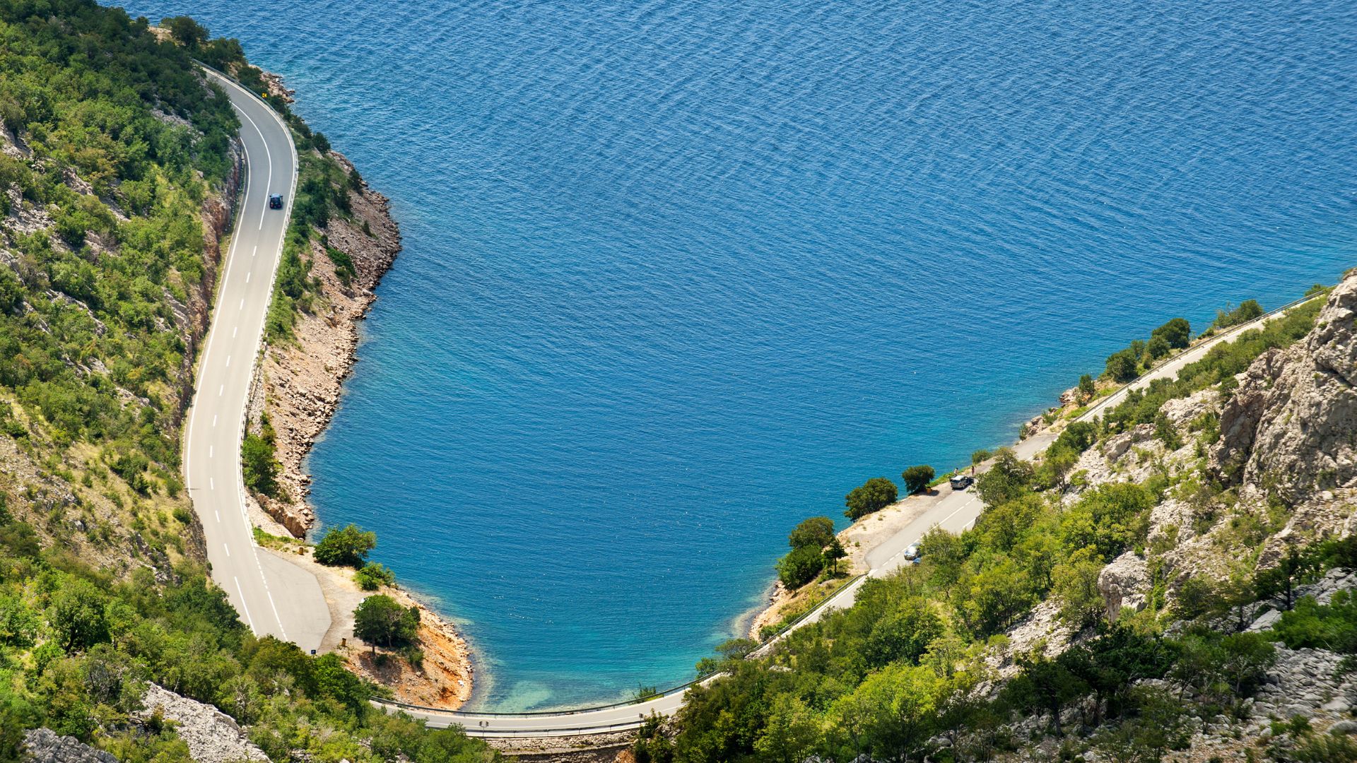 A high-angle view of a winding coastal road hugging a lush green hillside next to a deep blue bay or sea, with a few vehicles visible on the road.