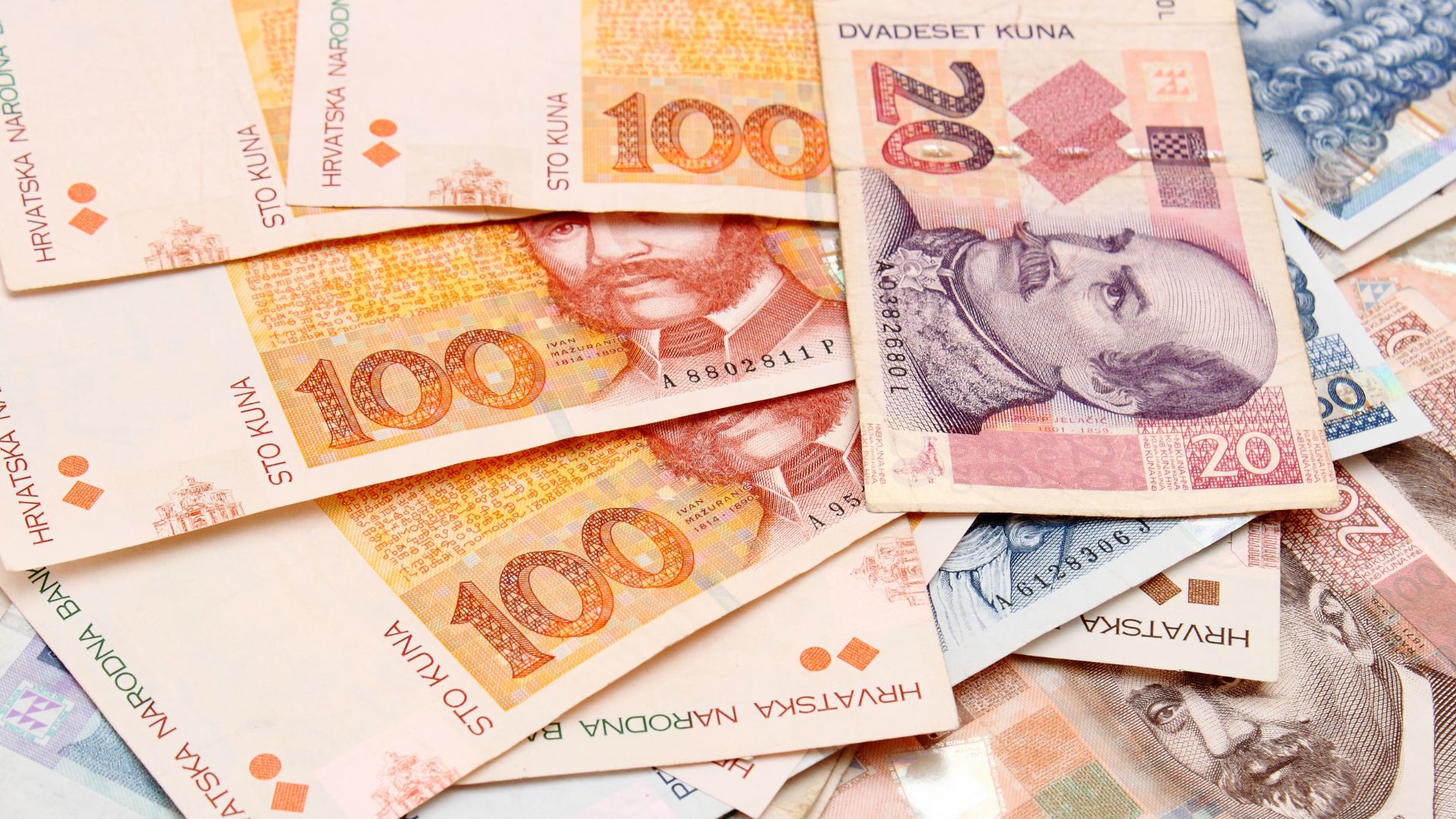 A close-up view of a scattered pile of Croatian Kuna banknotes, including 100 kuna notes featuring Ivan Mažuranić and a 20 kuna note featuring Josip Jelačić.