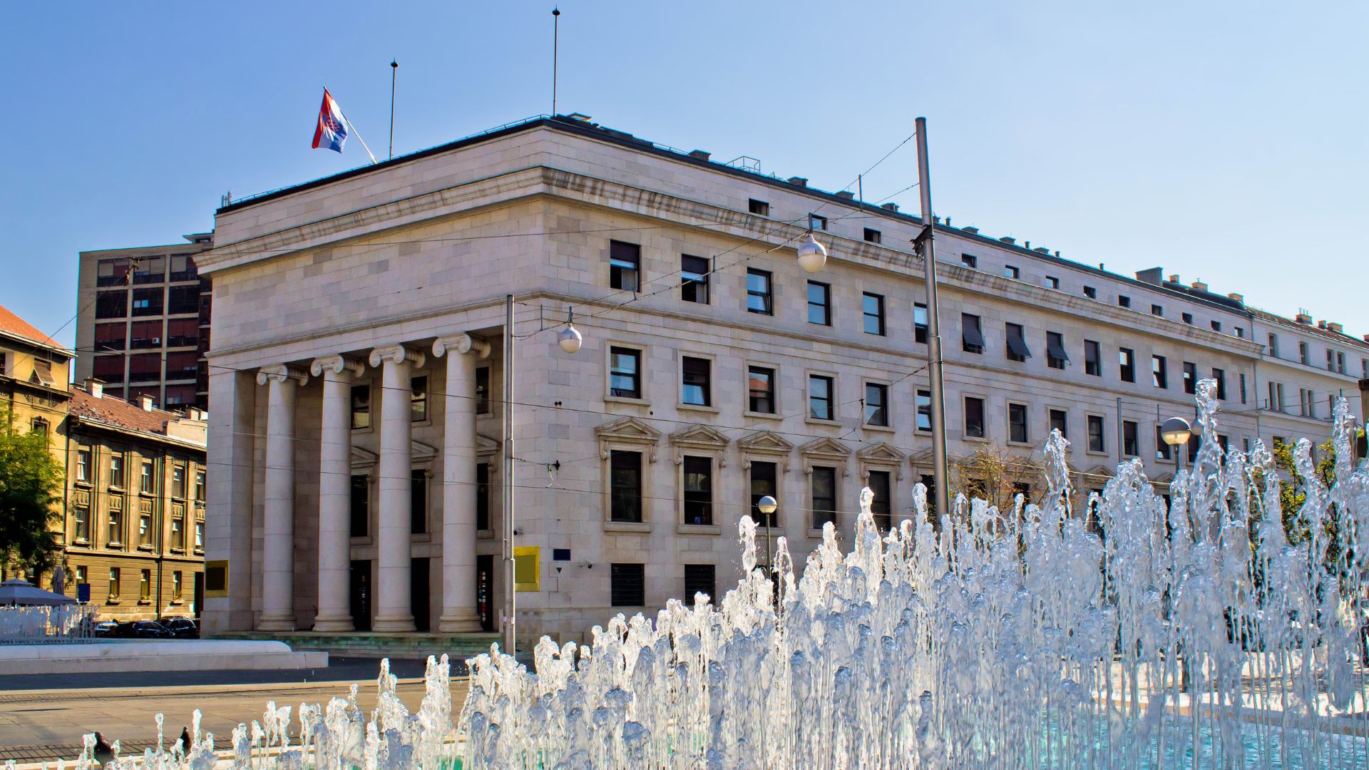 A neoclassical building with a prominent facade featuring four tall Ionic columns, fronting a city square with a fountain and a street with utility poles and traffic signals. The building is the Croatian National Bank in Zagreb.
