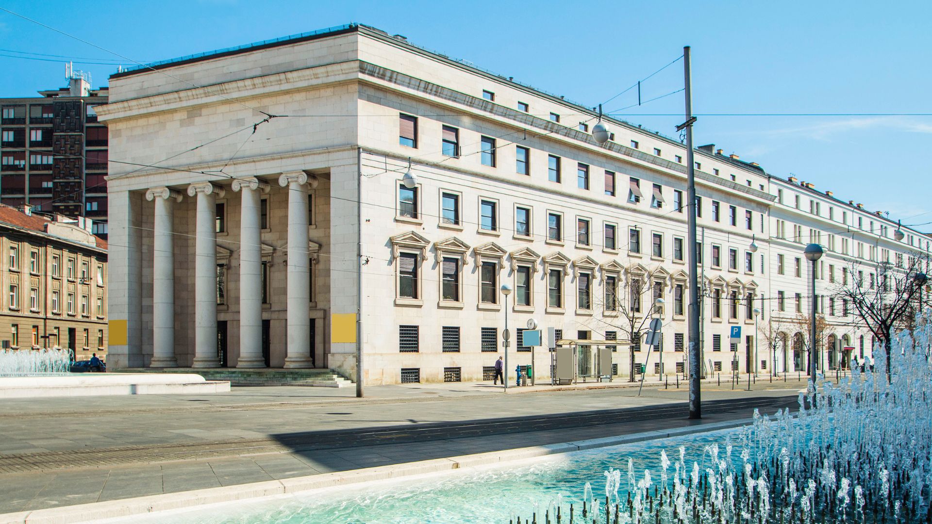 A neoclassical building with a prominent facade featuring four tall Ionic columns, fronting a city square with a fountain and a street with utility poles and traffic signals. The building is the Croatian National Bank in Zagreb.