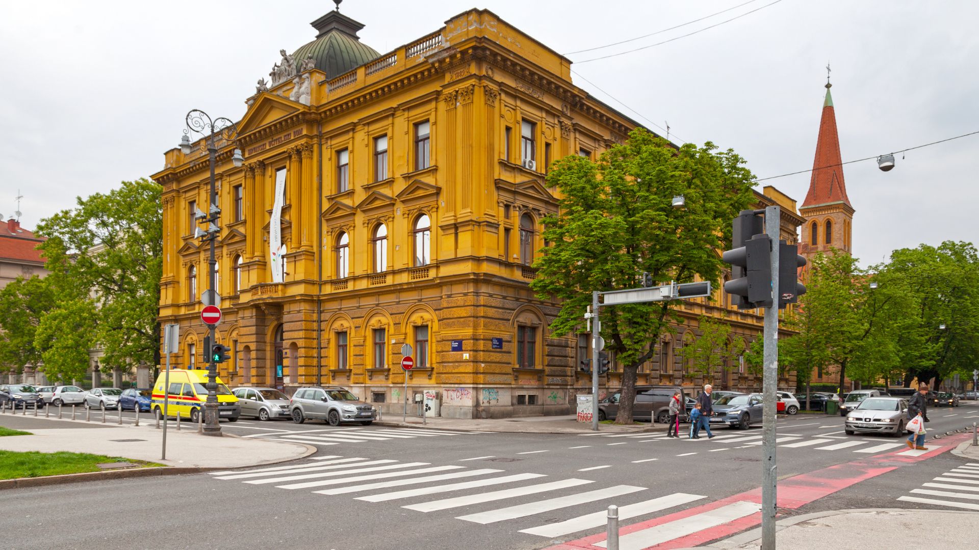 A grand, yellow, ornate building with a domed roof and multiple windows, identified as the Croatian School Museum, stands at a street intersection in Zagreb, Croatia. In the background, a red-spired church is visible, and the foreground features a crosswalk, traffic lights, and parked cars along the street.