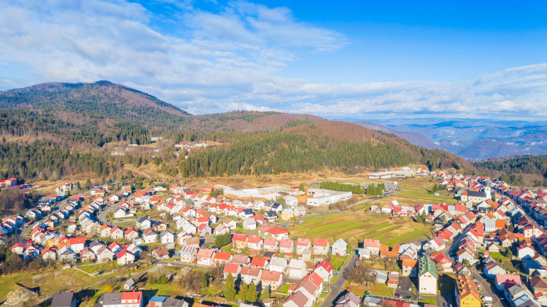 Aerial view of a town nestled in a valley with houses and buildings surrounded by forested hills and mountains under a blue sky with scattered clouds.