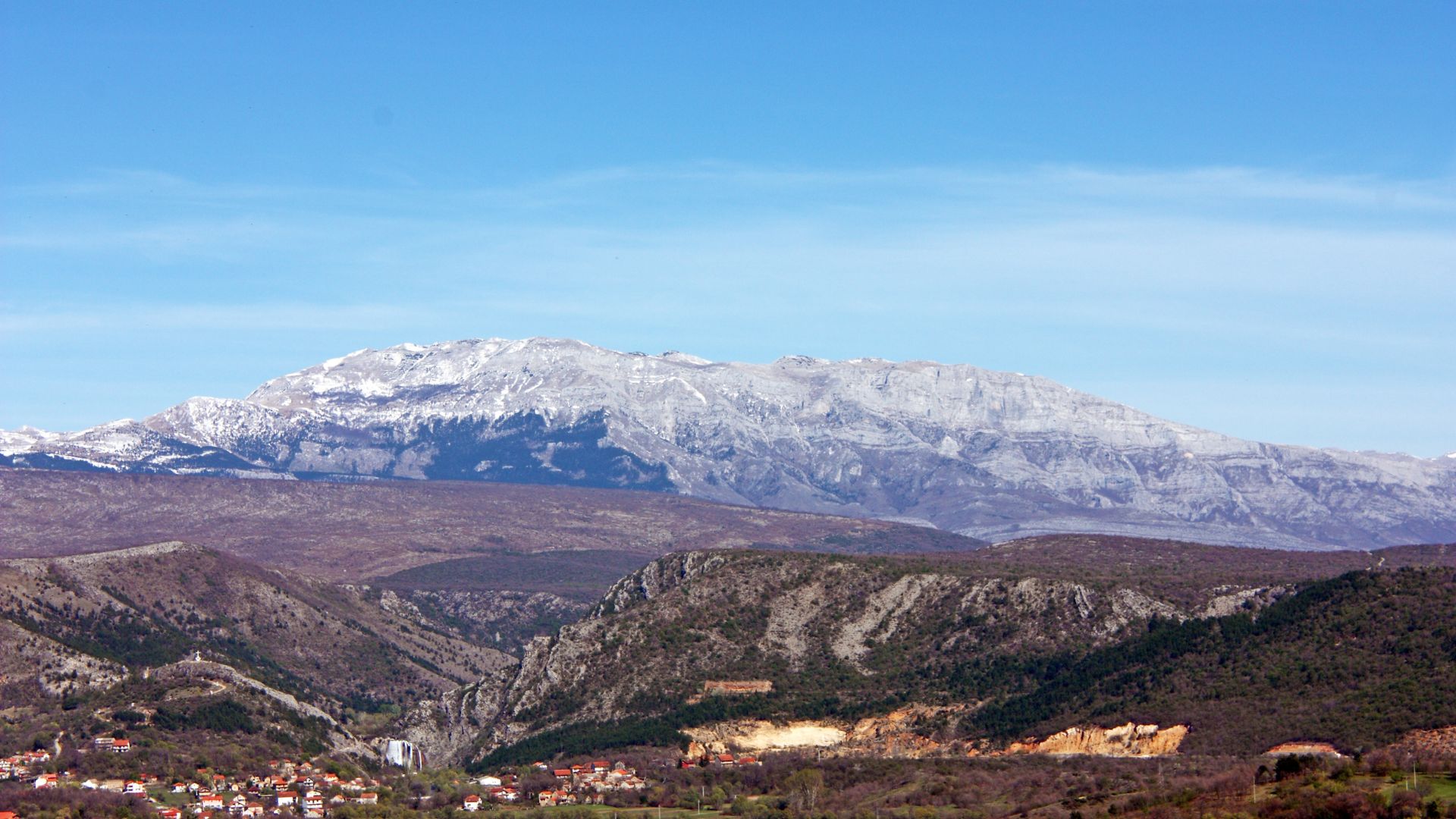 A wide-angle view of a rocky, dry landscape with sparse vegetation and a prominent mountain range under a clear blue sky.