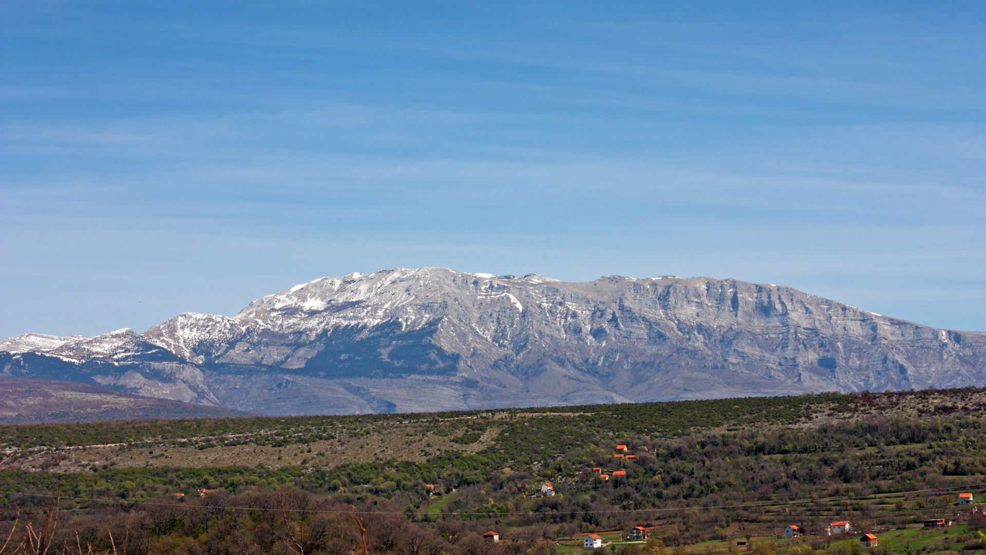 A panoramic view of the snow-capped Dinara mountain range under a clear blue sky, with a foreground of green and brown fields dotted with small houses and sparse trees.