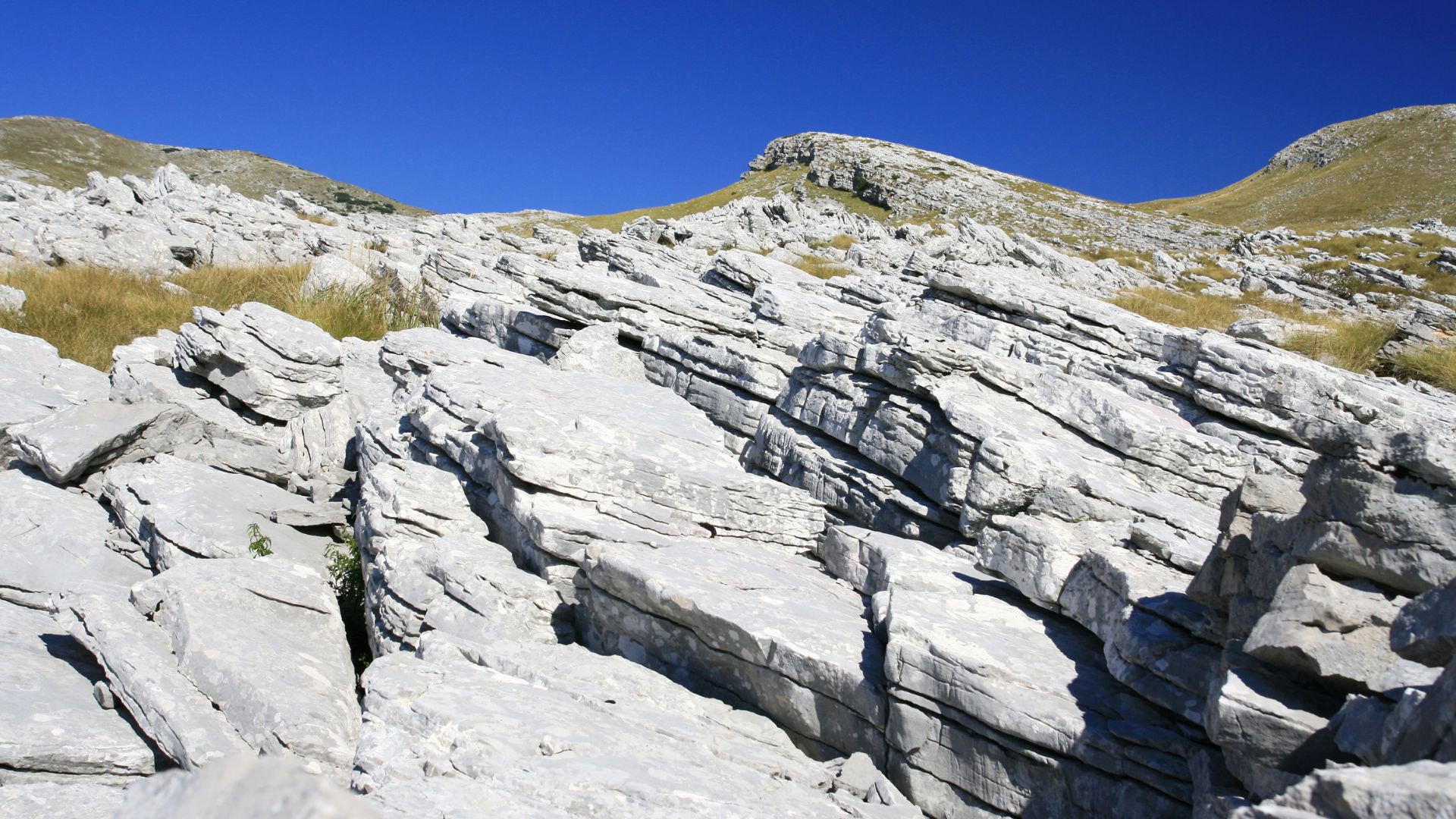 A wide-angle shot of rocky, layered white limestone formations on Dinara Mountain under a clear blue sky, with sparse patches of dry grass visible on the slopes.