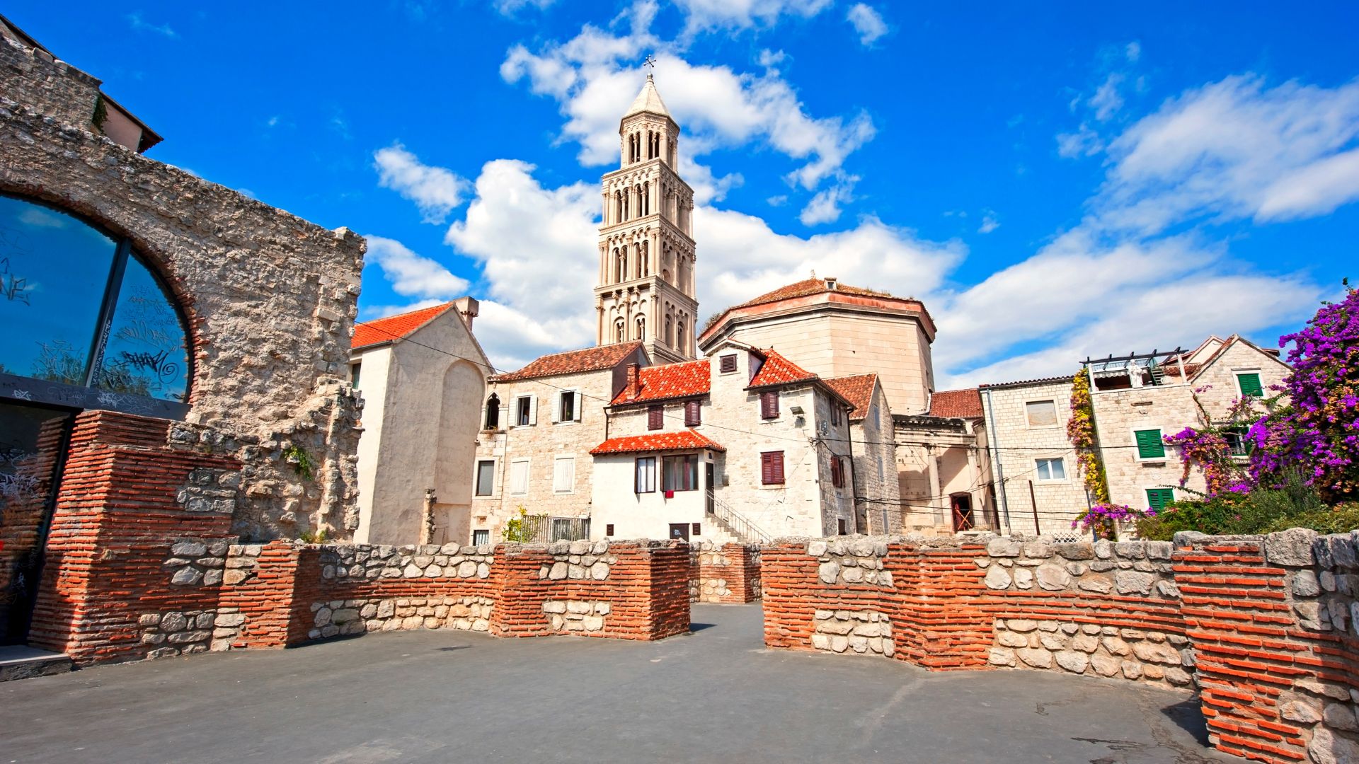 A low-angle shot captures the ancient Roman architecture of Diocletian's Palace in Split, Croatia, under a bright blue sky with scattered clouds. The scene features weathered stone walls and red-tiled roofs, with a prominent bell tower rising in the background. In the foreground, a paved area is framed by low stone and brick walls, leading towards the historic structures.