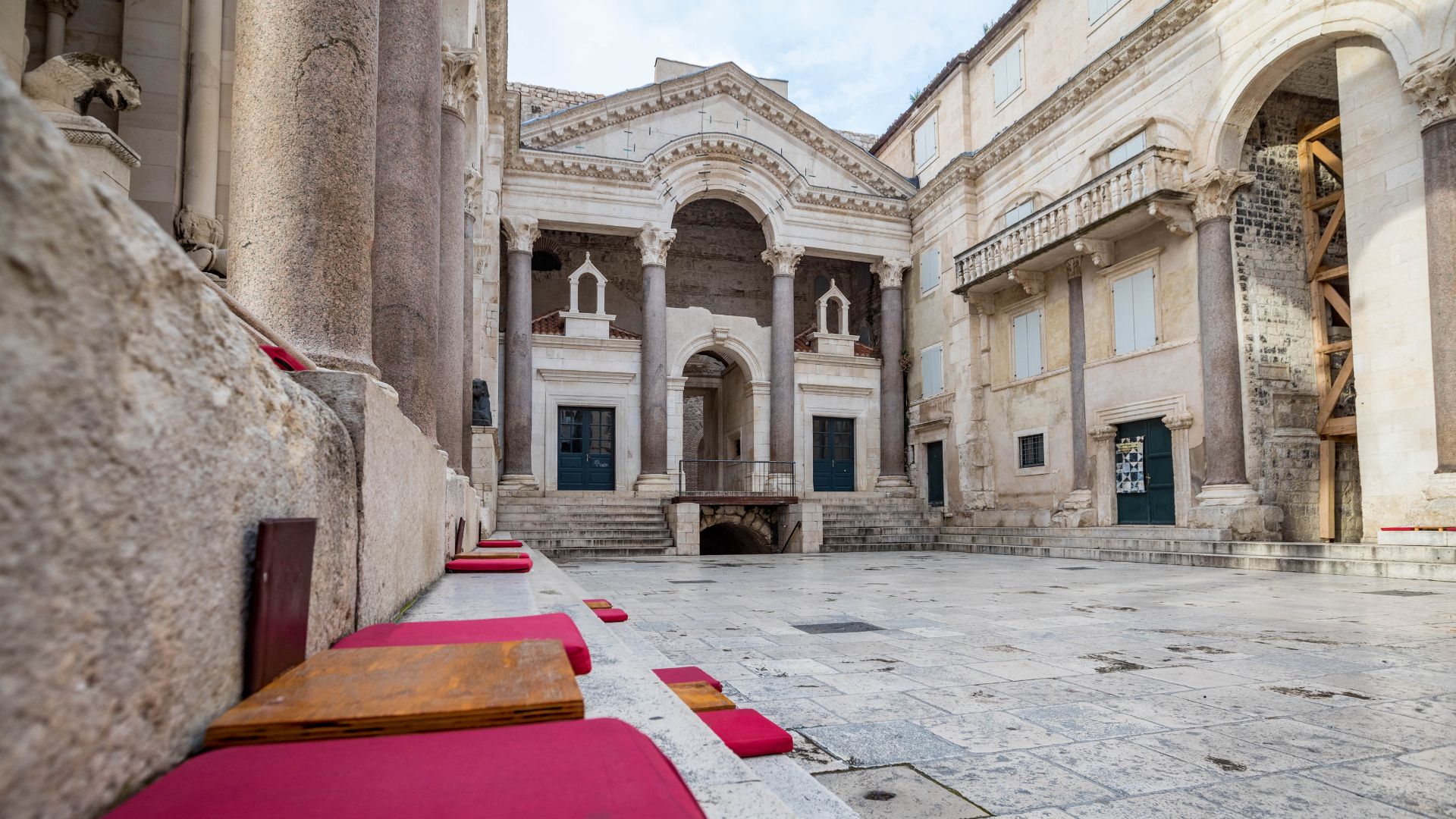 A low-angle shot captures the grandeur of Diocletian's Palace in Split, Croatia, showcasing its ancient Roman architecture. In the foreground, red cushions rest on stone steps, leading up to a grand entrance with towering columns and arches. The central courtyard is paved with stone, and surrounding buildings also exhibit classical architectural elements, including balconies and arched windows, under a bright sky.