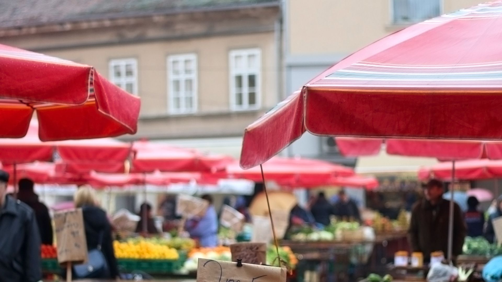 A bustling open-air market, Dolac Market in Zagreb, Croatia, is seen from a slightly elevated perspective. Numerous red umbrellas provide shade over stalls filled with fresh produce and other goods, while shoppers and vendors move among them. In the background, traditional European buildings with windows are visible.